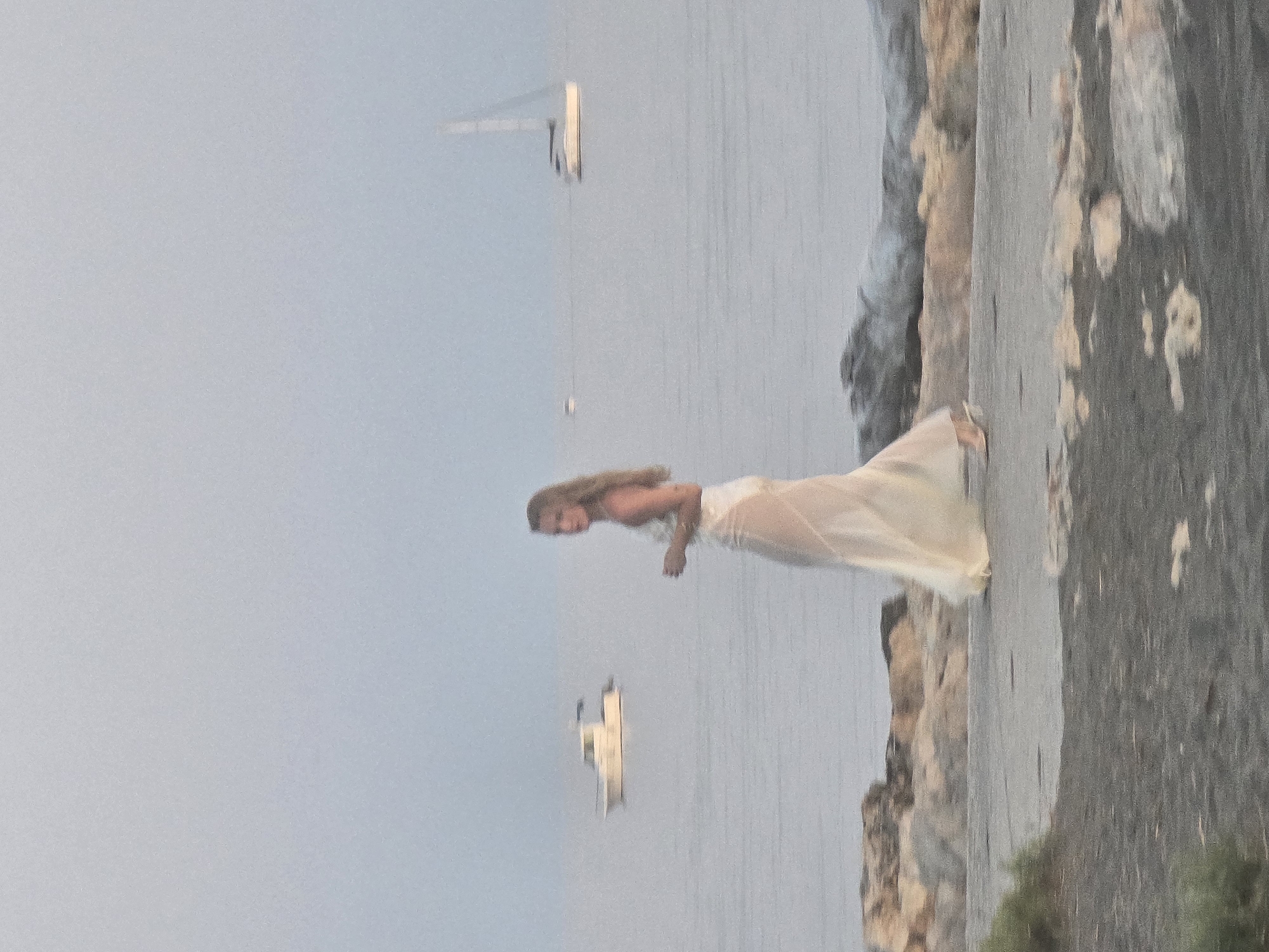 An adult female with blonde hair, wearing a light-colored, sheer dress, walks along a paved path next to a rocky shoreline. The sea is visible behind her, with a white boat in the mid-ground to her left and a sailboat in the background to her right. The sky is overcast. The scene is likely the Mediterranean coast near Málaga, Spain, given the style of the boat and the general coastal landscape.