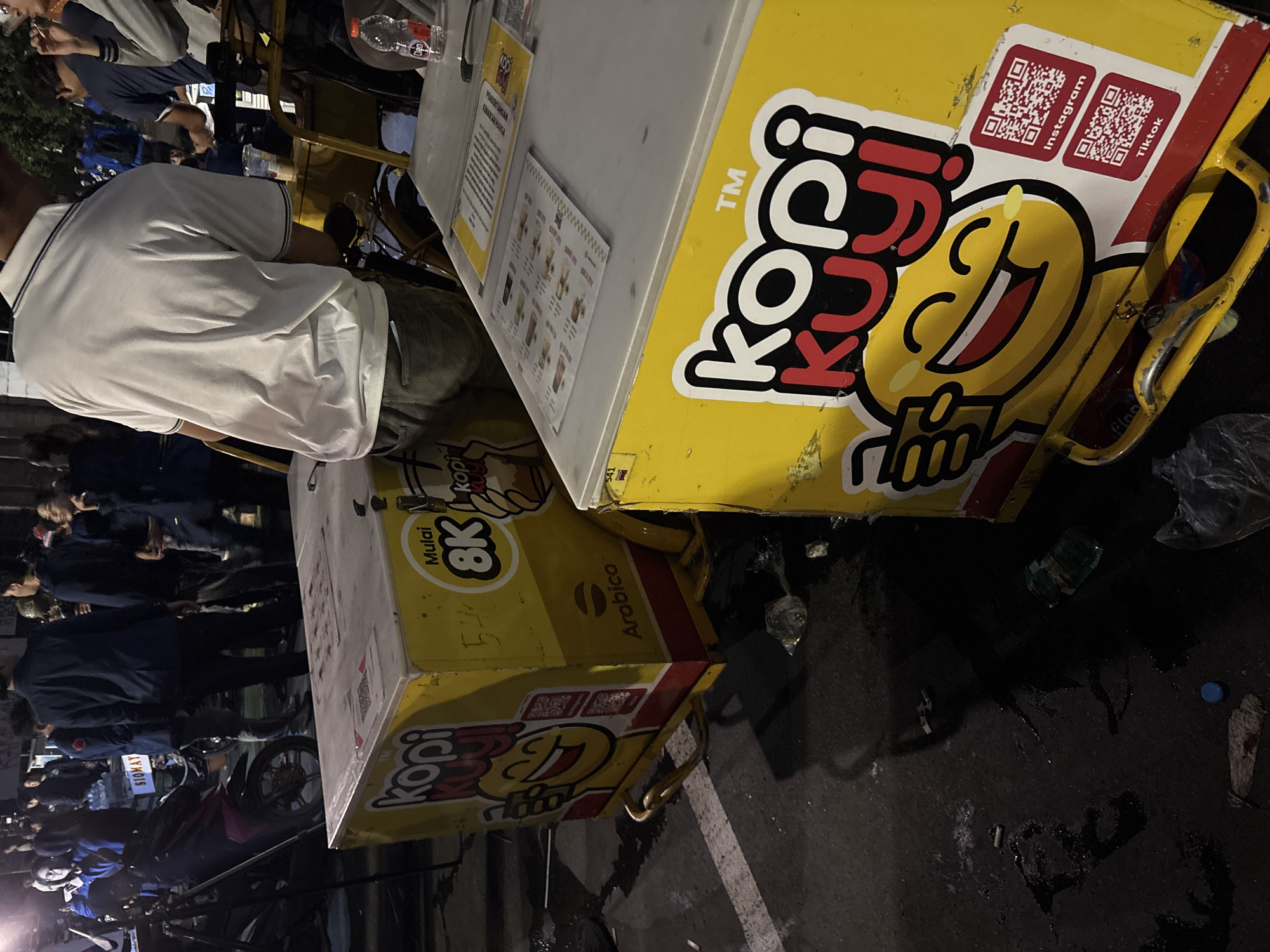 The image captures a street scene at night in Central Jakarta, Indonesia, likely during a bustling evening. The primary subjects are people and two prominent yellow food carts. One cart is emblazoned with the "Kopi Kuji" logo, featuring a smiling cartoon character and QR codes for Instagram and TikTok. The other cart, also yellow and featuring the "Kopi Kuji" branding, is positioned behind the first and appears to be serving a drink.
Several individuals are visible, some standing near the carts, possibly as customers or vendors, while others are blurred in the background, suggesting a crowded environment. One person in the foreground is wearing a white polo shirt and appears to be standing by a cart. The setting is a street with asphalt paving, and there are indications of a market or a popular gathering spot due to the presence of multiple food carts and people.
Notable details include the bright yellow of the food carts, which stands out against the dark surroundings of nighttime. The visible text on the carts, "Kopi Kuji," "Mulai 8K," "Arabica," "Instagram," and "TikTok," provides context about the product being sold. The presence of QR codes suggests a modern approach to marketing. The ground appears wet in places, and there is some litter, which is common in busy urban environments at night. The overall atmosphere seems lively and active, typical of a night market or a popular street food location.