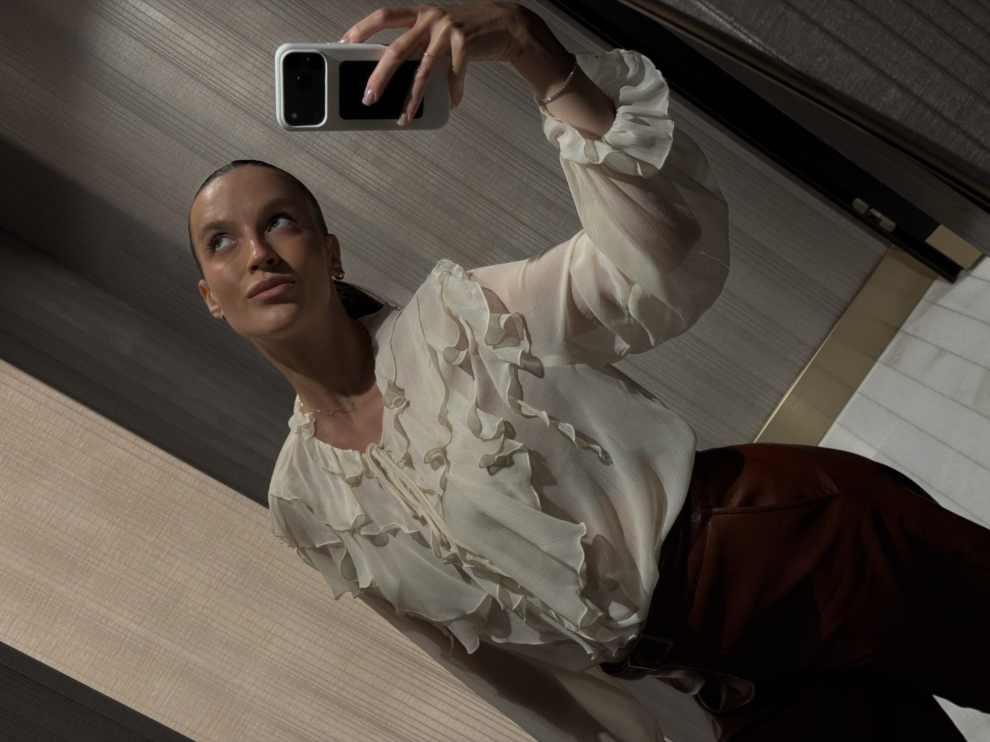 A woman is taking a mirror selfie in what appears to be a fitting room or dressing room. She is wearing a cream-colored, sheer blouse with ruffles and voluminous sleeves, and what look like dark red leather pants with a dark belt. She is holding a white phone in her right hand. The background consists of textured walls in shades of beige and dark grey, with some wooden paneling and a floor that looks like tiled or wooden planks. The lighting is dim and somewhat dramatic, creating shadows. The scene does not provide any specific visual cues to indicate the location is the city of Budva, Montenegro.