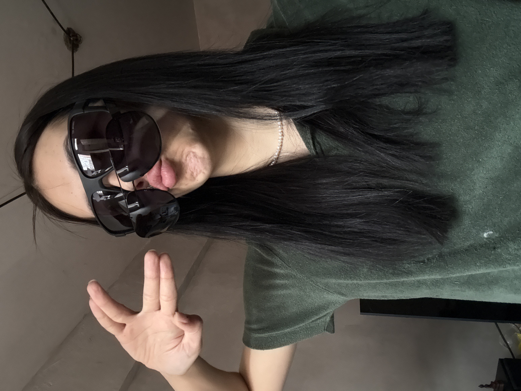 A person with long, black hair is wearing two pairs of sunglasses and sticking their tongue out slightly. They are wearing a green shirt and a pearl necklace. Their hand is raised with the ring and middle fingers together and the index and pinky fingers separated. The background includes a light-colored wall, a ceiling fixture, and a partially visible television. There is no information in the provided image to identify the location as Votkinsk, Russia.