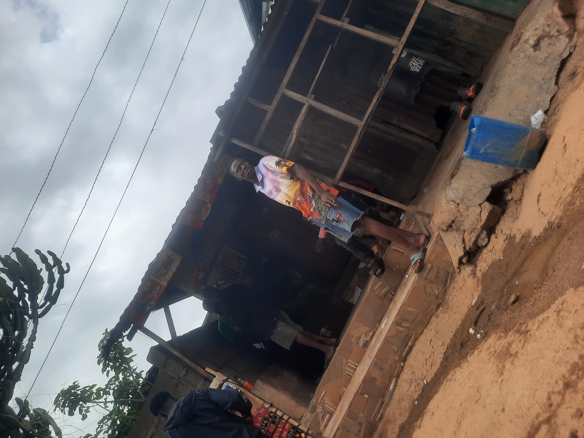 This street scene, likely in Gwong, Nigeria, depicts a small, rustic building, possibly a shop, under an overcast sky. The image is taken from a low angle, looking upwards towards the building and sky.
In the center, a young man poses for the camera, standing in front of the building's entrance. He wears a vibrant, colorful short-sleeved shirt featuring a landscape design with palm trees and a sunset, paired with distressed denim shorts and light-colored sandals that may have "OFF" visible on the strap. He looks directly at the viewer with a neutral expression.
The building itself appears to be constructed with a wooden frame and corrugated metal roofing, showing signs of wear. Under the eaves on the left, faded advertisements are visible, including text like "PEPSI" and possibly "LUCOZADE." The interior of the building is dark, but the faint outlines of other people can be seen inside. To the far left, another person, wearing a dark top, is partially visible, leaning over red crates stacked near the building. The crates have the word "KISS" printed on them, suggesting a brand of beverages.
The ground in the foreground is a reddish-brown dirt path, uneven and showing patches of wetness or small puddles, indicating either recent rain or poor drainage. A blue plastic jerrycan or container rests near the building's entrance on the right. Overhead, three power lines stretch across the grey, cloudy sky. On the far left, a tall, multi-branched plant with spiky or segmented stems, possibly a type of cactus or euphorbia, adds to the outdoor setting.