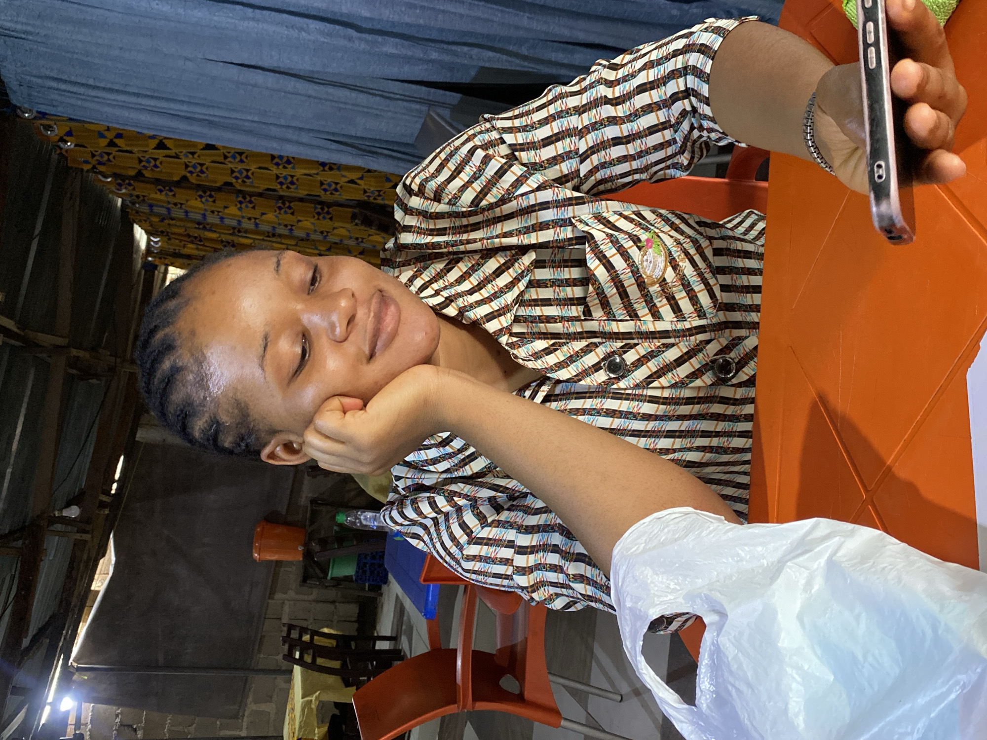 A woman with braided hair is sitting at an orange table, looking down at her phone. She is wearing a checkered shirt and has her hand resting on her cheek. Beside her is a white plastic bag. The background suggests an informal indoor setting, possibly a market or roadside eatery, with corrugated metal roofing, cinder block walls, and colorful curtains. Orange plastic chairs are visible in the background, along with some tables covered with yellow tablecloths. The overall scene conveys a casual and everyday moment. The presence of the plastic bag and the general ambiance suggest this might be in Jos, Nigeria, a city known for its vibrant informal markets and roadside food stalls.