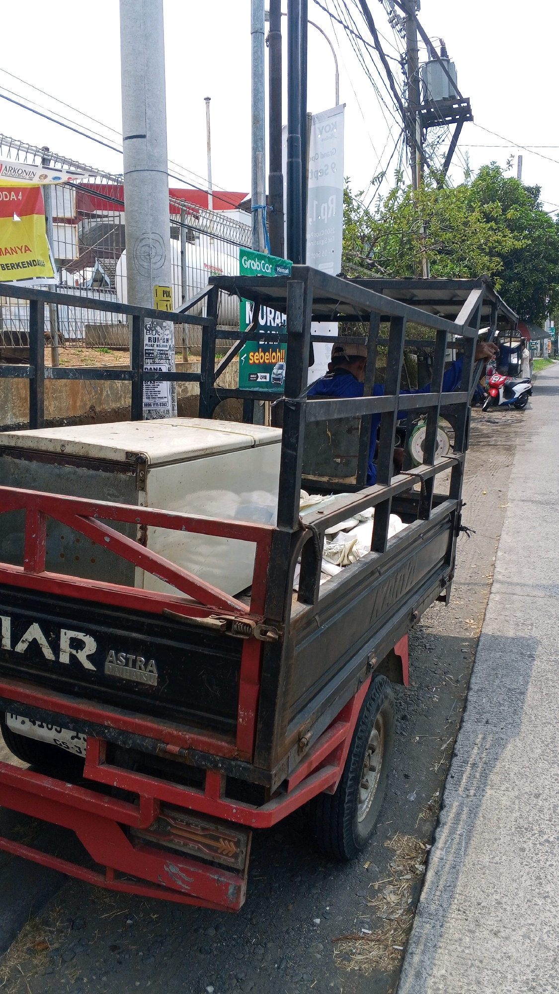 In the city of Semarang, Indonesia, a close-up shot captures a red and black utility truck with a metal cage. The truck is parked on a paved road, with a sidewalk bordering it and dry leaves scattered along the edge. In the bed of the truck, a white chest freezer is visible, along with some bundled white fabric. Two men, dressed in blue shirts, are seated within the truck's cage, appearing to be engaged in conversation or resting. One man wears a cap, while the other has his back to the camera. Behind the truck, a white utility pole stands tall, adorned with various signs and wires. Further in the background, green trees line the street, and a transformer is mounted on a wooden pole. Several motorcycles are parked along the side of the road. The overall scene is bathed in daylight, with a bright, overcast sky.