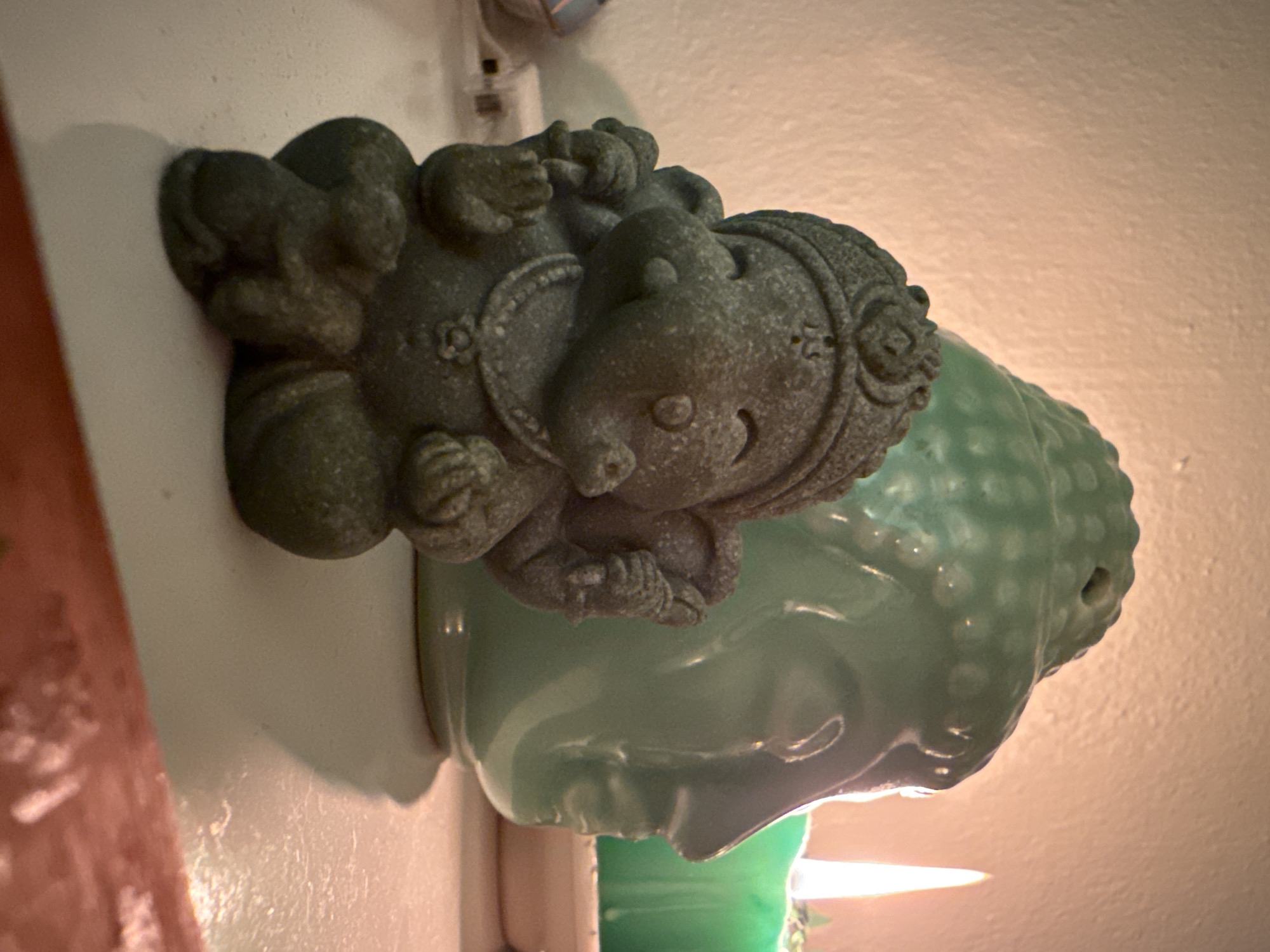 This is a medium shot, eye-level, indoor photograph of devotional objects illuminated by warm light. In the foreground, a small, dark gray, stone-like figurine of Ganesha, a Hindu deity, sits on a white surface. The Ganesha figurine is depicted with an elephant head, a smiling face, and is adorned with a headdress and necklace. Two small figures, possibly monkeys, are nestled at the base of the Ganesha figurine. Behind Ganesha, a large, light green ceramic Buddha head-shaped object is visible, with a textured surface and a hole on top, suggesting it may be a diffuser or incense burner. To the right of the Buddha head, a lit green candle burns, casting a warm glow. The background is a textured, cream-colored wall. The overall scene suggests a meditative or spiritual setting. The image does not provide any information about the city of Kadıköy, Turkey.