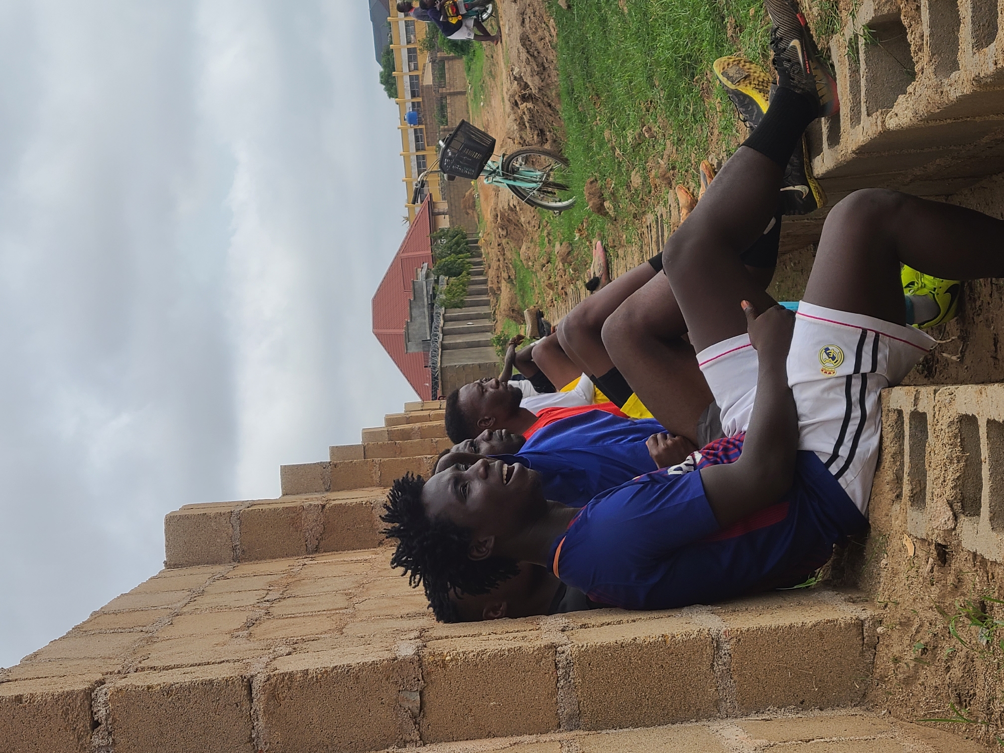 Several young men are seen resting outdoors on a stepped structure made of light-colored concrete blocks. There are at least five individuals visible. The closest man, with dreadlocks, is in a blue shirt and is smiling broadly, looking upwards. Others behind him are also reclining or lying down, dressed in casual sports attire, including t-shirts and shorts. One person wears white shorts with black stripes and a crest, possibly a sports team logo, on the leg. Athletic shoes, including a pair of black and yellow Nike sneakers, are also visible.
The scene is set outdoors under an overcast sky during daytime, suggesting diffused light. The background features a mix of dirt, sparse green grass, and distant buildings. A red-roofed building is present in the mid-ground, and a larger, lighter-colored multi-story building can be seen further away. A light blue bicycle with a black basket is parked upright in the dirt area behind the men. The overall mood appears relaxed and positive, particularly due to the smiling expression of the foreground subject.
