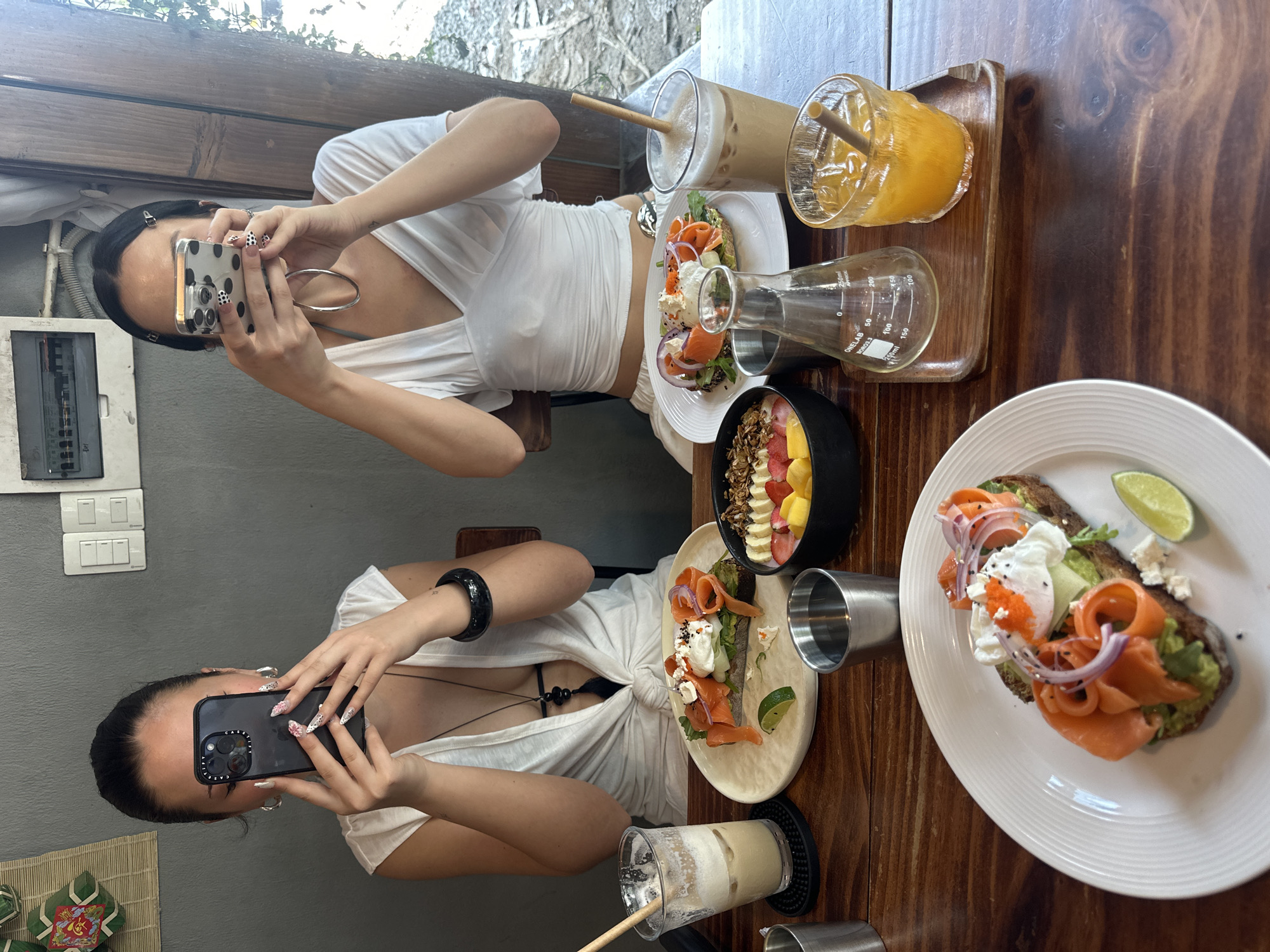 Two women are sitting at a wooden table in a restaurant. One woman is taking a picture of the other, who is posing with a similar action. Both women are wearing white shirts. The table is set with food and drinks, including a smoothie bowl, avocado toast with salmon, and various beverages. The location appears to be Đà Nẵng, Vietnam, based on prior context. Electrical and wall-mounted objects are on the left.
