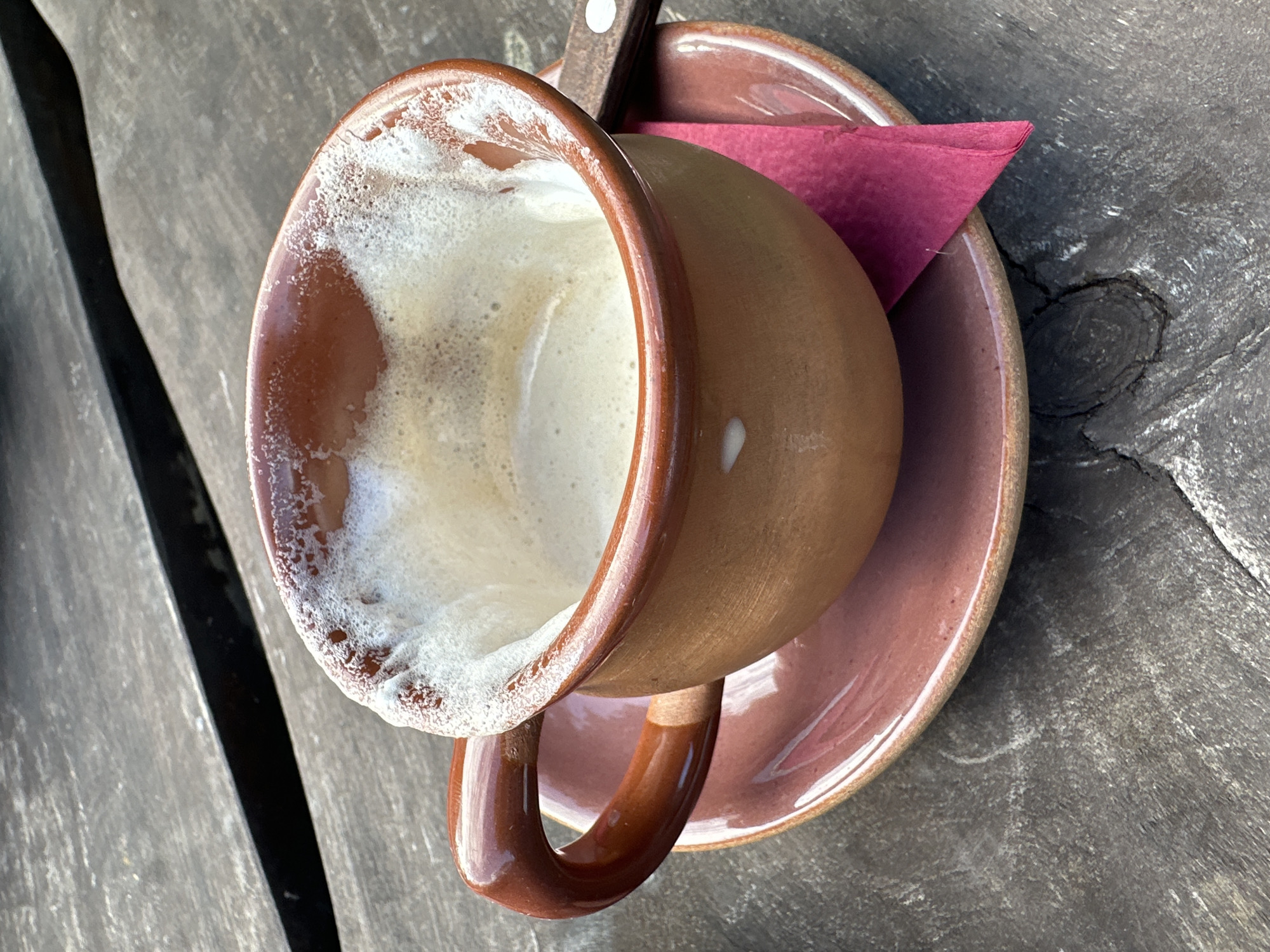 A close-up, high-angle shot shows a ceramic coffee cup and saucer on a rustic wooden table. The cup, made of terracotta-colored ceramic with a glossy brown glaze on the handle and rim, contains a foamy, light-brown beverage, suggesting it's nearly empty. A dark pink napkin is tucked beneath the saucer, peeking out from under the cup. The wooden table has a weathered, distressed finish with visible grain and cracks. The scene appears to be outdoors due to the natural lighting. This image is not located in Saula, Estonia.