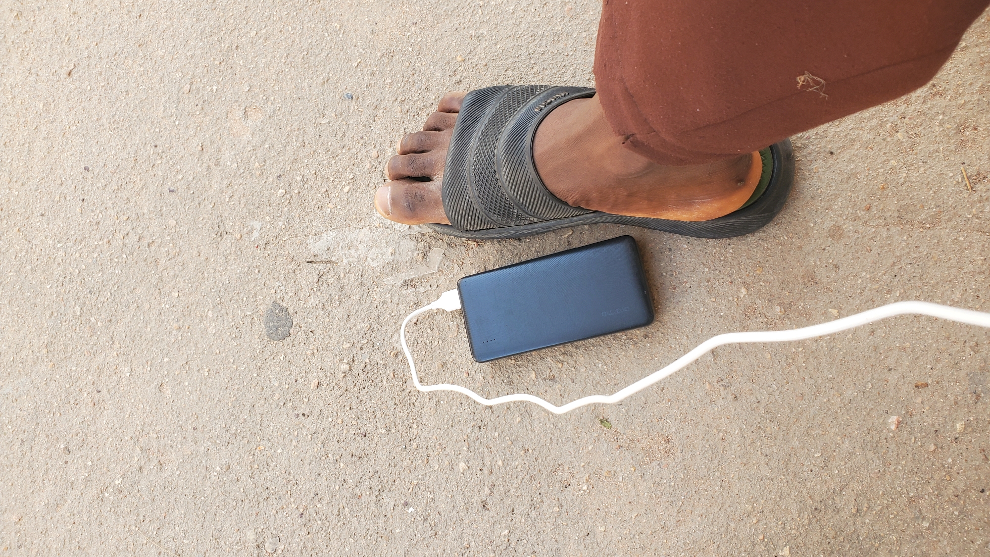 An overhead shot shows a black power bank connected to a white cable, resting on a granular, light brown asphalt surface. To the right of the power bank is a bare foot wearing a black sandal, with the toes pointing upwards. The foot is positioned on the right side of the frame, and a portion of brown trousers is visible on the right side of the image. The asphalt surface appears to be outdoors, suggesting a public space such as a street or pavement within the city of Jalingo, Nigeria. There are small stones and a few dried plant fragments scattered across the ground. The lighting suggests it is daytime.
