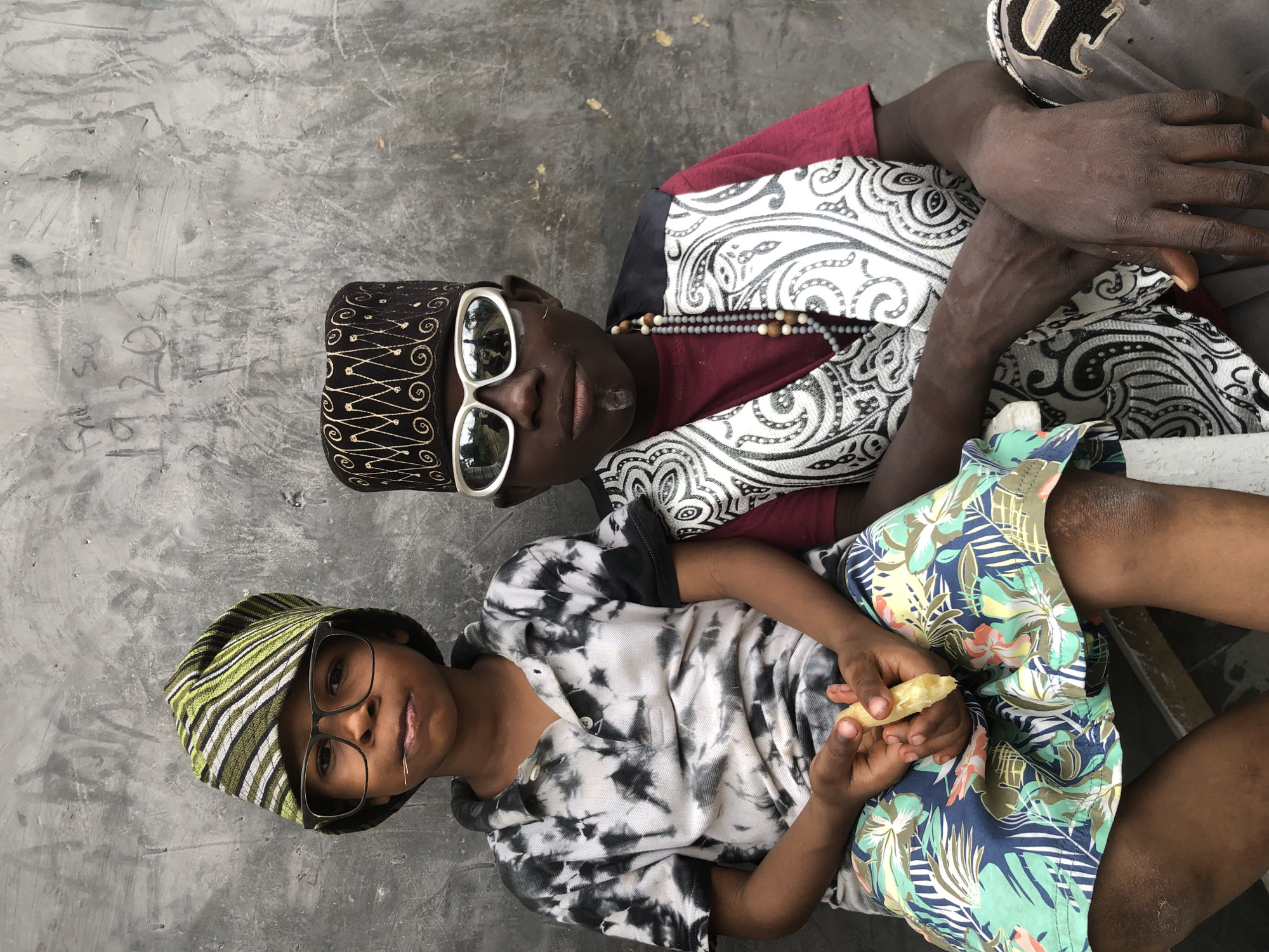 Two young boys are seated in front of a rough, textured concrete wall in Ado, Nigeria. The boy on the left wears a tie-dye polo shirt and a green and black striped cap. He has glasses on and is holding a piece of yellow food in his hands. The boy on the right wears a maroon t-shirt, a patterned vest, a black cap with gold embellishments, and large white sunglasses. He has a beaded necklace and a small white mark on his chin. The wall behind them is gray and has some faint writing or markings on it. The lighting suggests it is daytime.