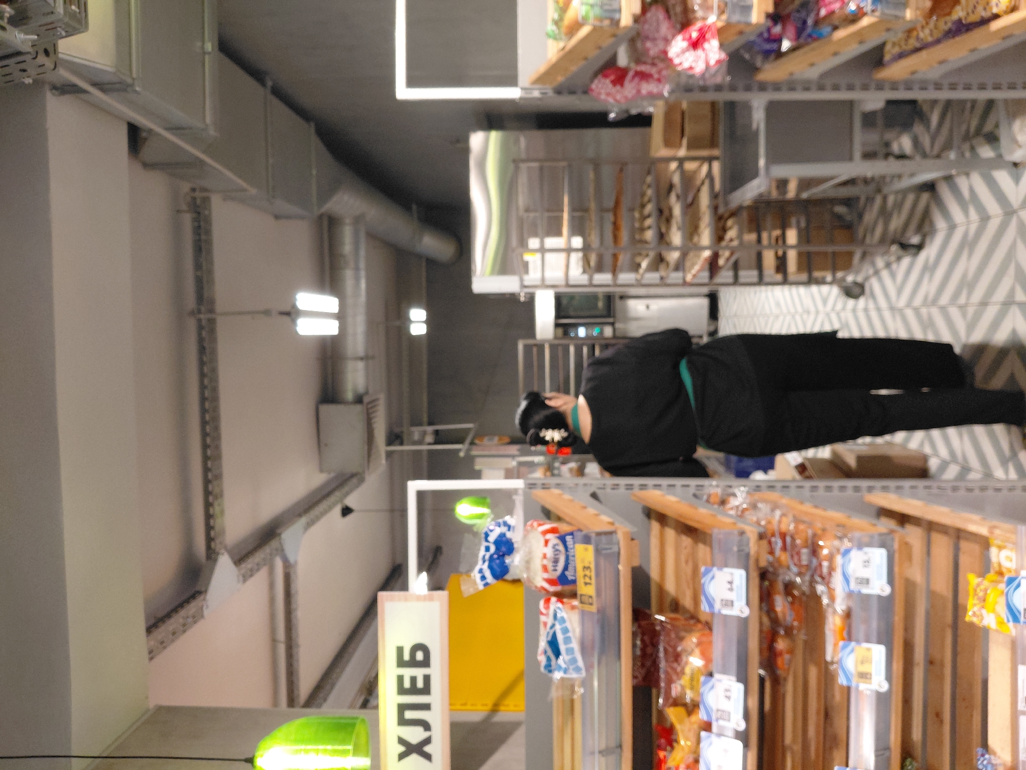 A medium shot captures a woman from behind, walking away from the camera in what appears to be a bakery or grocery store aisle. She wears a black uniform with a green apron. The aisle is lined with shelves displaying bread and baked goods. A sign above the shelves reads "ХЛЕБ", which translates to "BREAD" in Russian. The floor has a distinctive chevron pattern. The setting is likely a commercial establishment in Novosibirsk, Russia, given the Cyrillic signage.