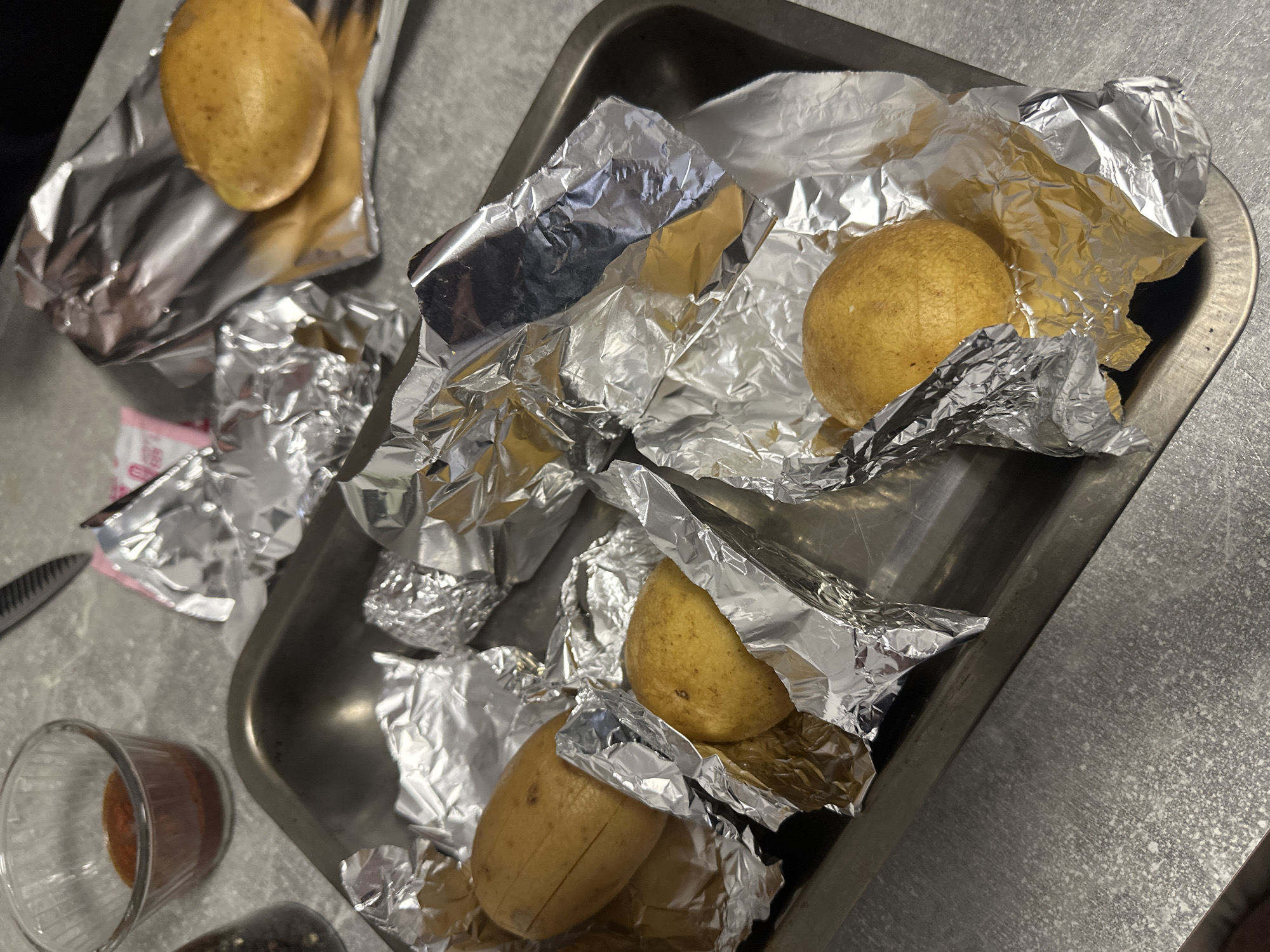 The image depicts a rectangular aluminum baking pan containing potatoes individually wrapped in aluminum foil. One potato is sliced in half. Some of the foil wrappers are open, revealing the potatoes. Next to the pan on a gray countertop, there's a glass containing a reddish-brown liquid, a knife, and what appears to be a grinder.
There is no specific information that allows me to conclusively determine the location is Varna, Bulgaria.