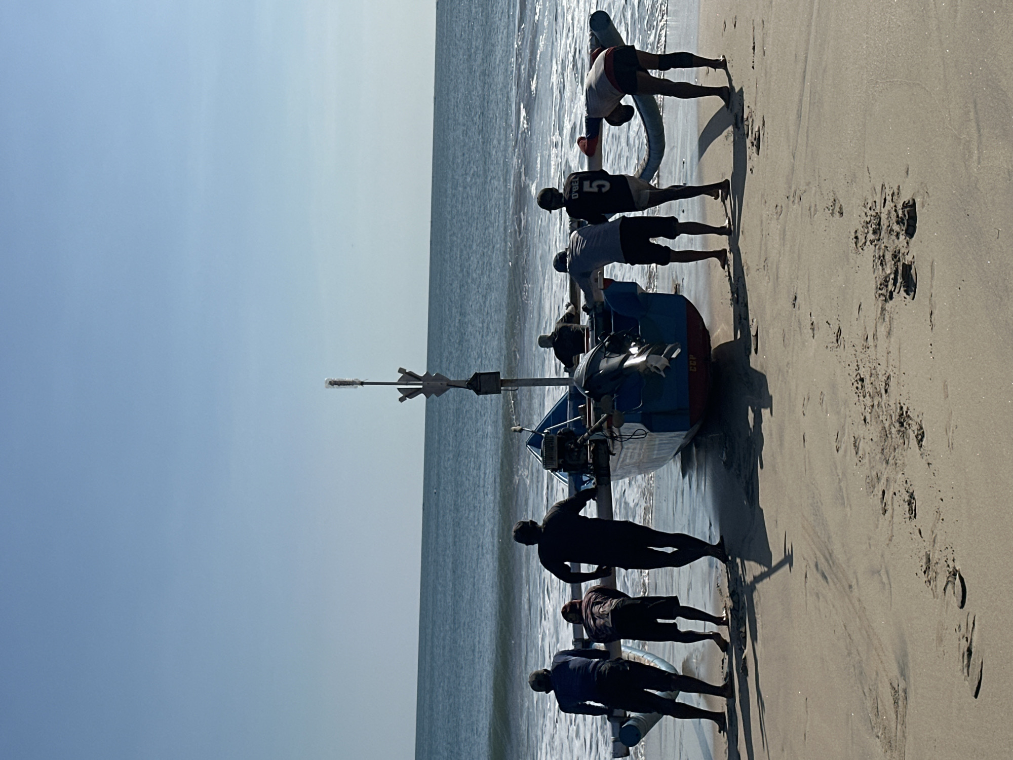 Eight individuals are on a sandy beach, pushing a blue and red fishing boat towards the ocean. The sky is clear and blue, with the ocean in the background showing moderate waves. The people are dressed casually, with some wearing shorts and t-shirts. Two of the individuals are wearing sports jerseys, one with the number "5" on the back. The boat has an outboard motor and a tall mast with a light at the top. Footprints are visible in the sand around the boat and people. The scene appears to be set during daylight hours. The location is the beach in Kuta, Indonesia.