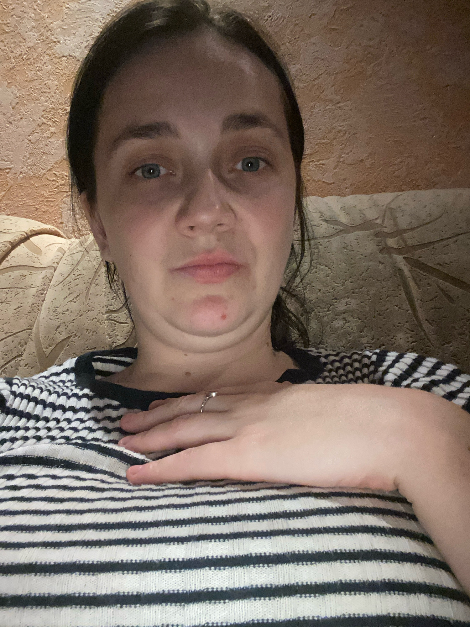 A woman is shown in a close-up, selfie-style shot. She has dark hair and blue eyes and is wearing a black and white striped shirt. Her left hand is resting on her chest, and she is wearing a ring on her index finger. The background appears to be a textured wall and a patterned couch. The lighting suggests an indoor setting, possibly at night. The overall mood is casual and intimate. The image does not provide any information about the city of Unknown, Unknown or any specific location details.