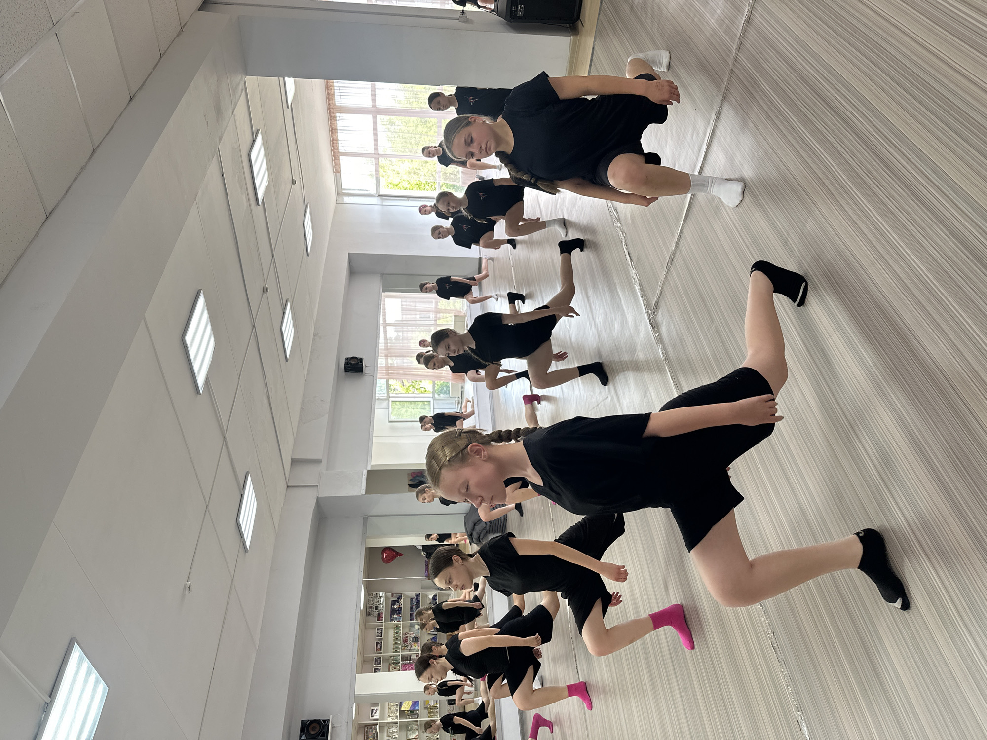In a dance studio, possibly located in Votkinsk, Russia, a group of young girls in black athletic wear are engaged in a stretching exercise, specifically a lunge. The studio features large mirrors on one wall, reflecting rows of trophies and awards on shelves. The floor is light-colored with a striped pattern, and the room is illuminated by fluorescent ceiling lights. The girls are positioned in various stages of the lunge, with their legs extended and bent. Some are wearing colorful socks, such as pink and white, while others are wearing black socks. The overall atmosphere suggests a dance class or training session.