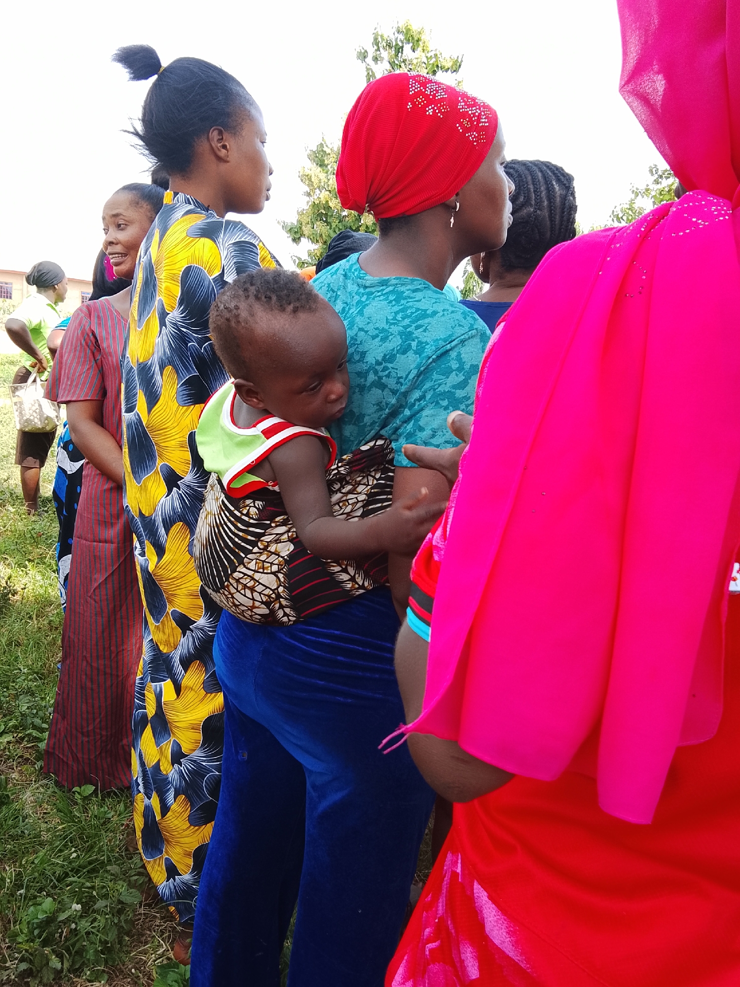 In Akwanga, Nigeria, a group of people stand outdoors on a grassy area. The foreground is dominated by a child being carried on the back of a woman wearing a bright blue velvety fabric covering her legs. The child, with dark skin and hair, is wrapped in a patterned fabric. The woman carrying the child is wearing a teal-colored shirt with a textured pattern. To her left, another woman is visible, wearing a vibrant yellow and blue patterned dress. Further to the left, a woman in a striped red and brown dress stands with a white bag in her hand. In the background, other individuals are present, and trees and a building can be seen. The overall scene suggests an outdoor gathering or event.