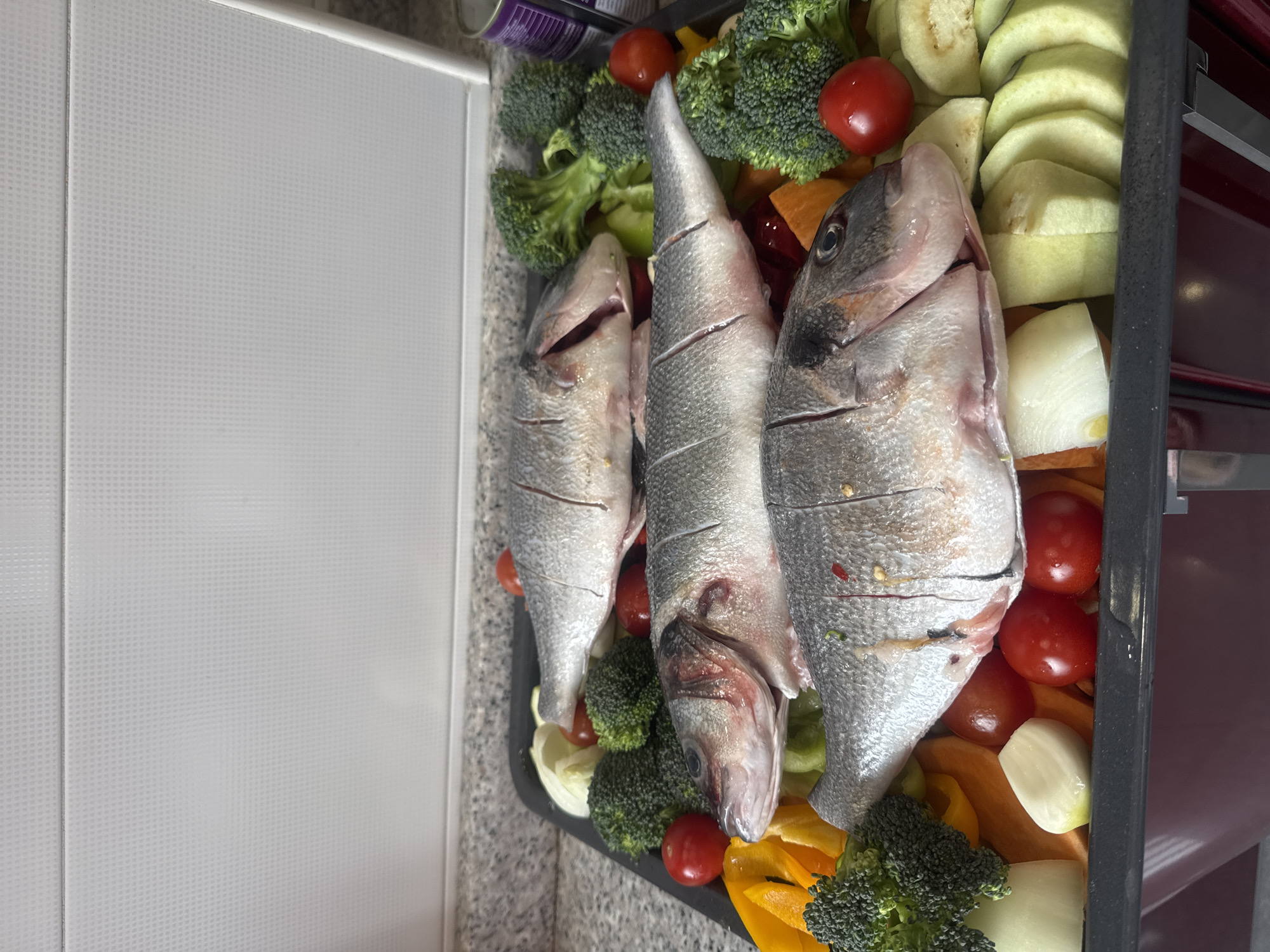 A prepared baking tray with three gutted fish surrounded by vegetables is being readied for cooking indoors. The fish are laid from left to right, and the vegetables include broccoli florets, cherry tomatoes, sliced zucchini, sliced onions, and yellow bell pepper pieces. The baking tray sits inside an oven frame. The picture was taken on a light countertop or table next to a white cabinet. There is no evidence the picture was taken in Tbilisi, Georgia.
