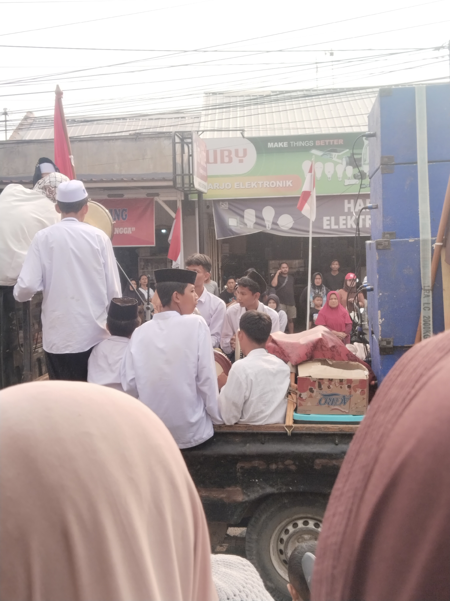 The image depicts a street scene in Balapulang Kulon, Indonesia. In the foreground, partially visible, are the heads of two individuals wearing headscarves, one in a light pink and the other in a reddish-brown hue. The middle ground is dominated by a flatbed truck, where several young boys in white clothing and skullcaps are seated. Some of them are holding or playing drums. Behind them, other people are standing or sitting.
On the left side of the truck, a young boy in a white shirt and black skullcap is positioned closer to the front. To his right, another boy in similar attire is visible. Further behind them, a taller individual in a white shirt and black skullcap is also seated on the truck. To the left of the truck, a person in a white robe and a white skullcap is standing, holding a red flag.
In the background, several storefronts are visible. One banner has text that reads "ARJO ELEKTRONIK" and another has "MAKE THINGS BETTER". There are also posters and advertisements displayed. On the far right of the image, a large blue speaker cabinet is positioned. The overall atmosphere suggests a local event or procession. The sky is overcast.