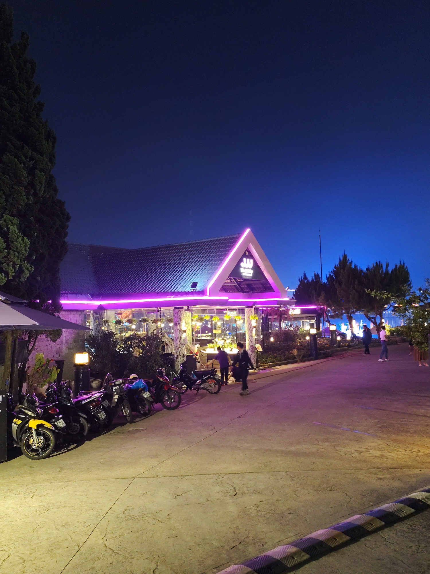 The image shows a building with a dark tiled roof and triangular entrance highlighted with pink neon lights, likely a restaurant or cafe, located in Da Lat, Vietnam. The surrounding area includes paved surfaces, trees, and parked motorcycles. Several people are seen walking or standing near the building. The sky is dark blue, suggesting it is either night or dusk.