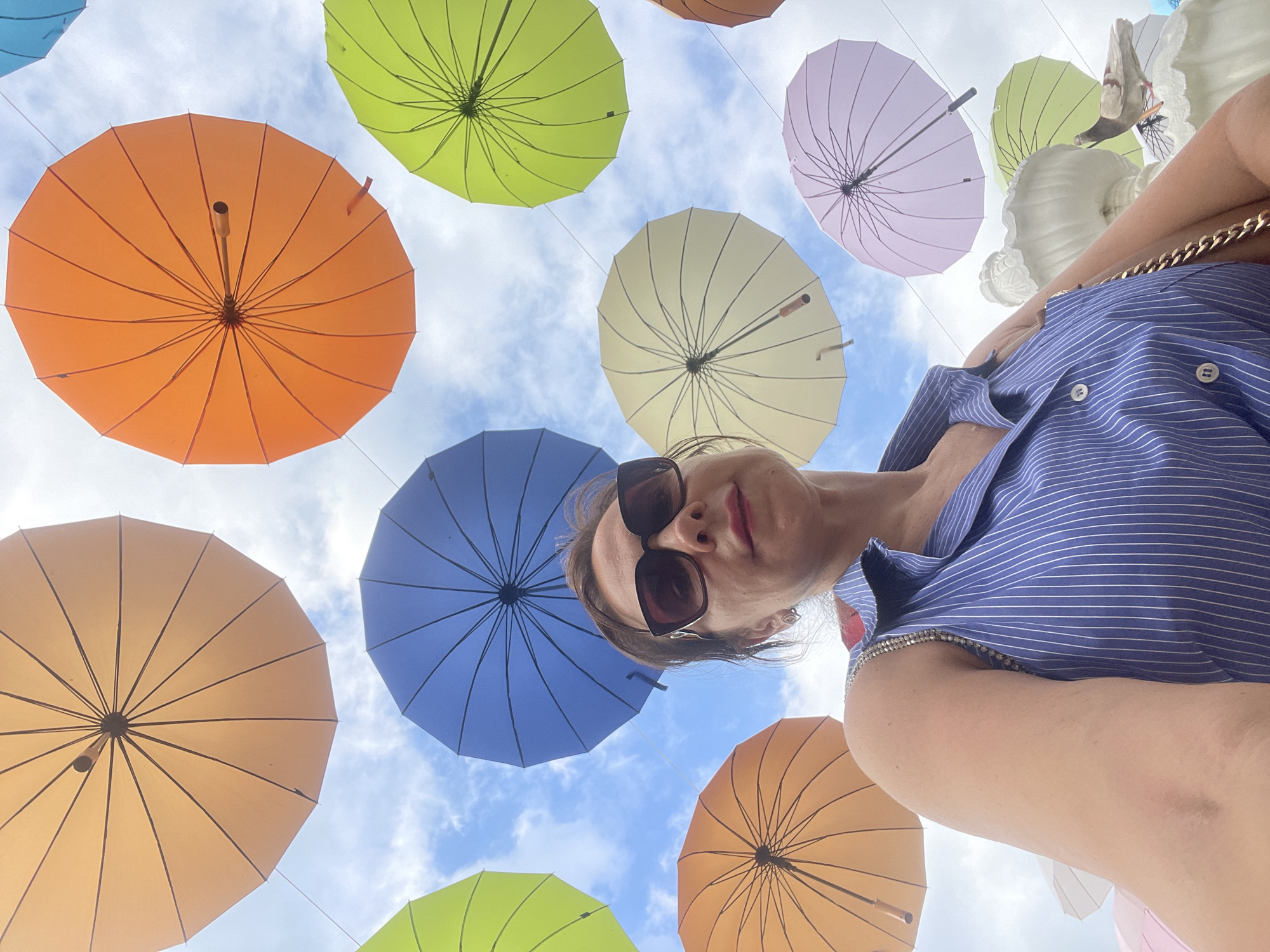 An adult female is taking a selfie outdoors with multiple colorful umbrellas suspended in the sky above her. The umbrellas are in various colors including orange, yellow, blue, lime green, pale yellow, and lavender. The woman is wearing a blue and white pinstripe sleeveless shirt with silver embellishments on the shoulder straps. She has on large dark sunglasses and is looking at the camera. The sky is visible through the umbrellas and appears to be partly cloudy. The location is identified as the Sochi Discovery World Aquarium in Sochi, Russia, which is known for its display of suspended umbrellas. In the lower right corner of the image, a pigeon is perched on a white decorative element, possibly part of the aquarium's landscaping.