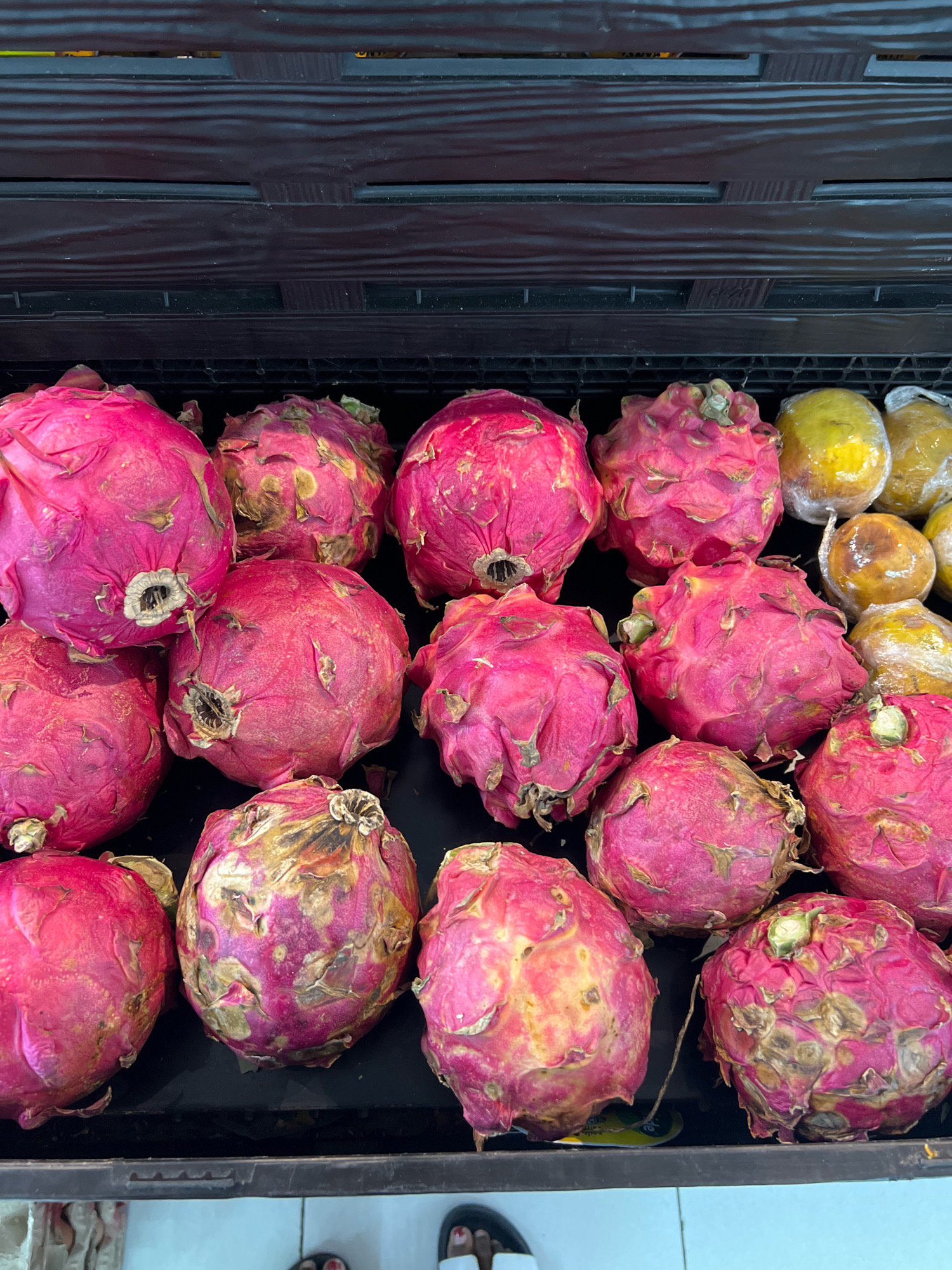 In Denpasar, Indonesia, a close-up, eye-level shot captures a display of pink dragon fruits and a few yellow fruits, possibly papayas, in a black plastic crate. The crate is filled with the fruits, with the dragon fruits being the dominant subject. The dragon fruits are various shades of pink, some with dried, brown, or yellowing skin and distinct green stems or buds. Some of the dragon fruits appear slightly bruised or weathered. To the right, several yellow fruits, wrapped in clear plastic, are visible. The background is a dark, textured surface, likely the interior of a market stall or refrigerator unit. The overall lighting is even, highlighting the textures and colors of the produce. No people are visible in the frame.