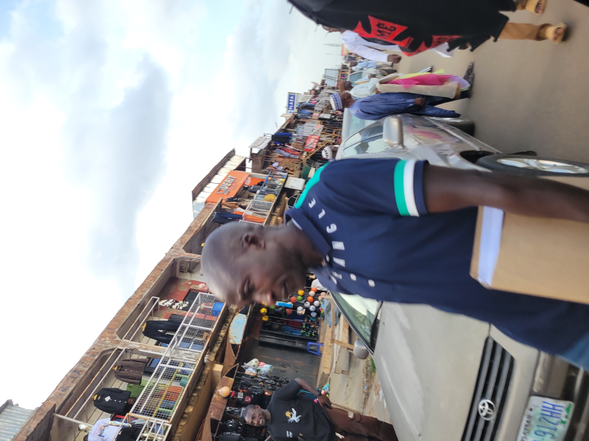 The image depicts a bustling street scene in Jos, Nigeria, likely during the daytime with an overcast sky. A group of people are navigating a crowded marketplace situated between buildings filled with merchandise. The foreground is dominated by a man wearing a dark blue t-shirt with green and white trim on the collar and sleeves. He is holding a light brown cardboard box. To his right, a silver sedan is parked, its front grille and license plate visible. The license plate reads "912HH".
Behind the man and the car, the market unfolds. Buildings lining the street are packed with shops, displaying clothing and other goods. Stalls are set up along the pathway, further narrowing the space for pedestrians. The overall atmosphere appears lively and busy, suggesting ongoing commercial activity. Various individuals, some carrying bags or parcels, are interspersed throughout the scene, contributing to the sense of a vibrant market day. The architecture of the buildings suggests a somewhat informal and densely packed urban environment.