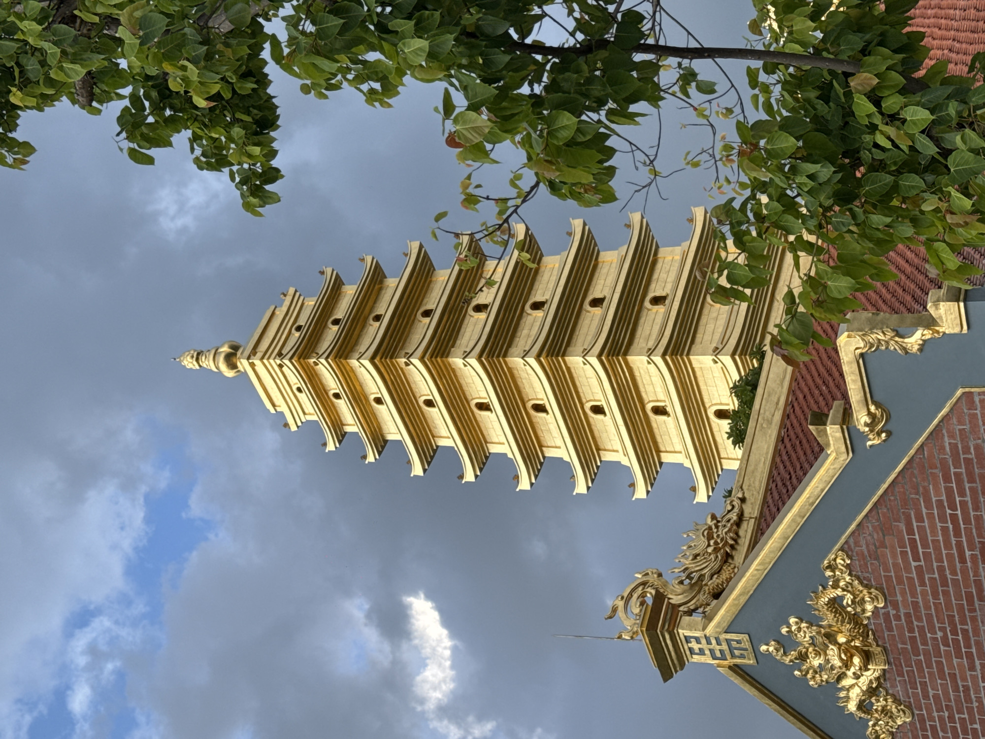The image depicts a golden pagoda in Diên Khánh, Vietnam, set against a cloudy sky. The pagoda is a multi-tiered structure with each level featuring curved roofs and small arched windows. A portion of a building with a red tiled roof and intricate golden dragon decorations is visible in the foreground, to the left of the pagoda. Lush green foliage from a tree partially obscures the right side of the pagoda and extends into the upper right corner of the frame. The lighting suggests it may be late afternoon or early evening, with the golden structure catching the sunlight. There are no people visible in the image.
