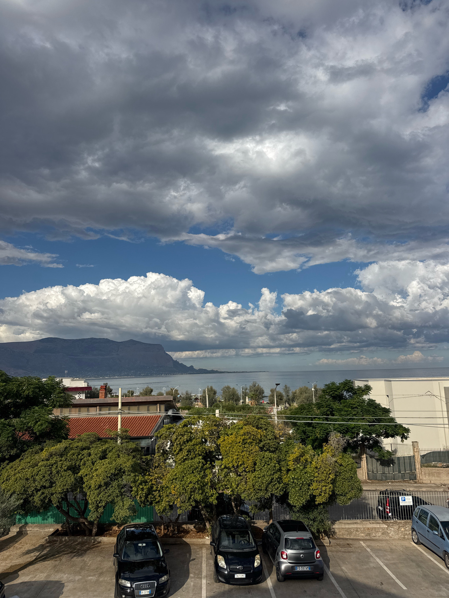 The image captures a view overlooking a parking lot and a body of water with mountains in the distance, likely in the city of Capaci, Italy. In the foreground, several cars are parked on a paved lot. A black Audi is parked on the left, followed by a silver Smart car, and a light blue car on the right. Behind the parking lot, lush green trees partially obscure a few buildings with red tile roofs. Beyond the trees and buildings, a calm body of water stretches towards a distant mountain range under a dramatic sky filled with both dark storm clouds and bright white cumulus clouds. The overall scene suggests a coastal area with variable weather conditions.