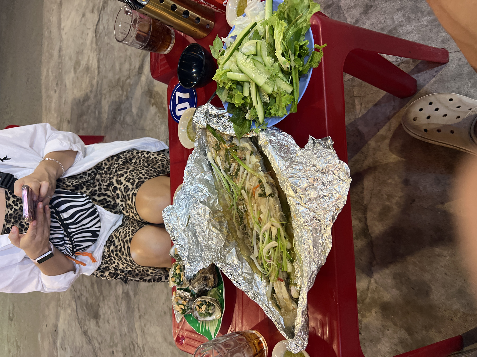The image depicts a top-down view of a scene likely in Bac Nha Trang Ward, Vietnam, given the context. A person is seated, dressed in a white shirt and animal-print shorts, holding a phone. A red plastic table contains several items: a blue plate of lettuce and cucumber slices, what appears to be foil-wrapped fish, a small dish of oysters, a glass of liquid, and a portable stove. Part of another person's leg and a Croc-style shoe are visible on the right side of the frame. The flooring appears to be concrete.
