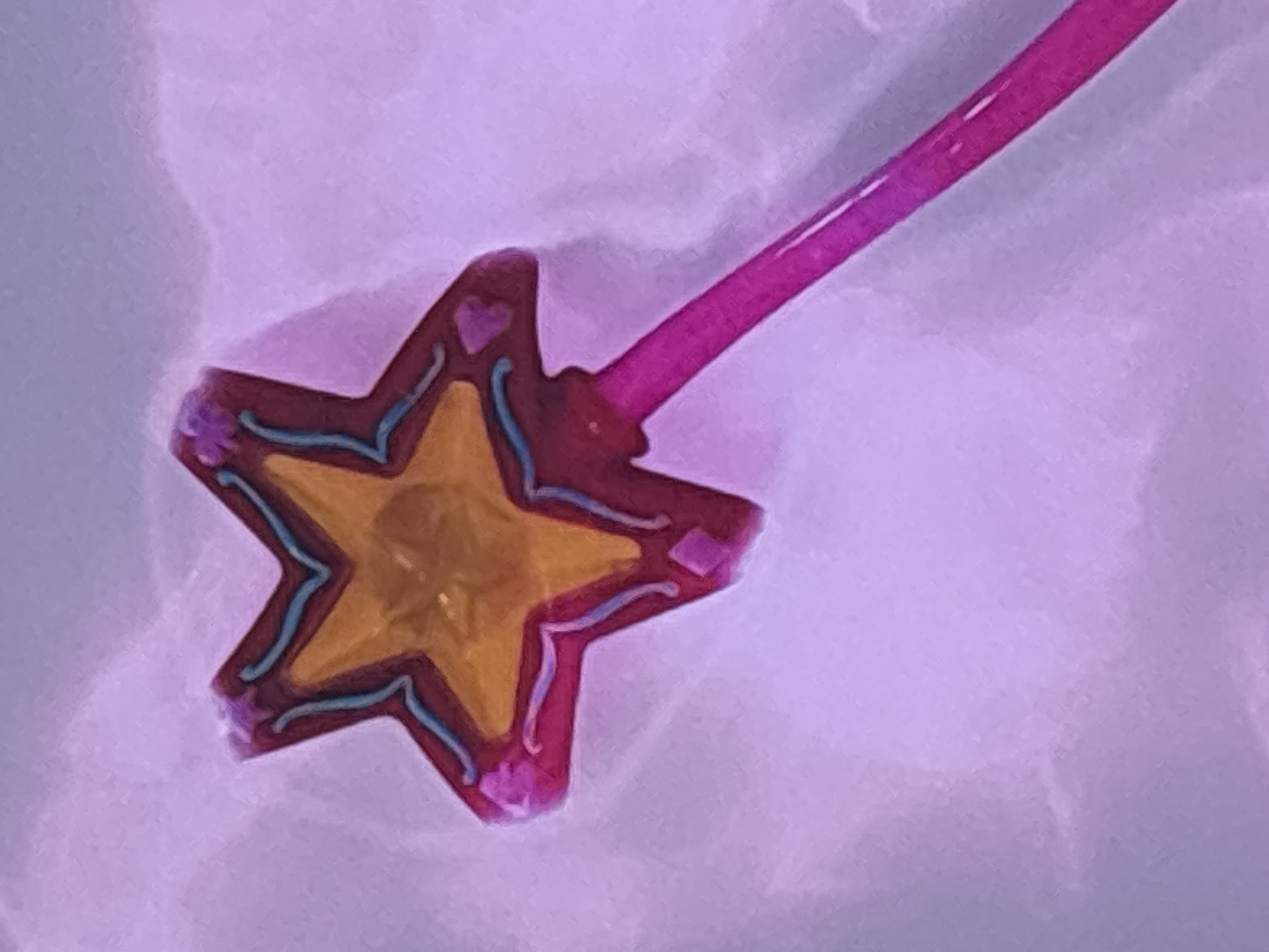 A close-up, overhead shot shows a pink plastic wand with a star-shaped tip against a blurred lavender background. The star is outlined in dark pink and decorated with teal lines and small purple flower shapes. The wand is positioned diagonally from the lower right to the upper left of the frame. There are no people or signs of life. The location is not discernible from the image.