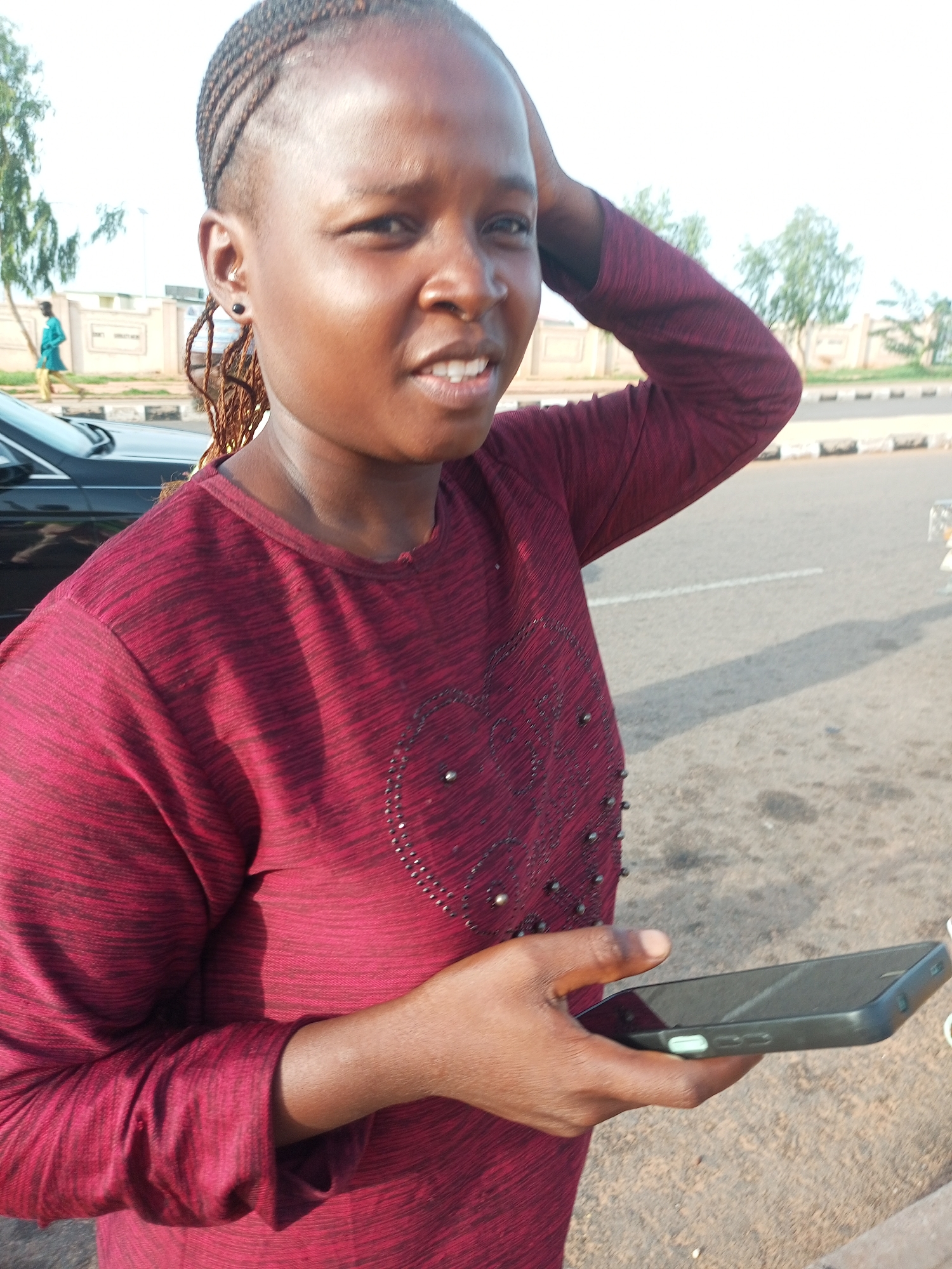 A medium shot shows a young Black woman with braided hair, standing outdoors in what appears to be a city. She is wearing a dark red long-sleeved shirt with a patterned texture and a heart design on the chest adorned with beads. The woman is holding a dark-colored smartphone in her right hand, which is positioned in front of her. Her left arm is raised and her hand is touching her head. Her expression is neutral, with her mouth slightly open. Behind her, a black car is partially visible on the left, and a road with white markings is in the background. Trees and a building are also visible in the distance. The lighting suggests it is daytime, with sunlight casting shadows on the ground. The ground is paved with asphalt and some gravel. The overall scene is unremarkable.
