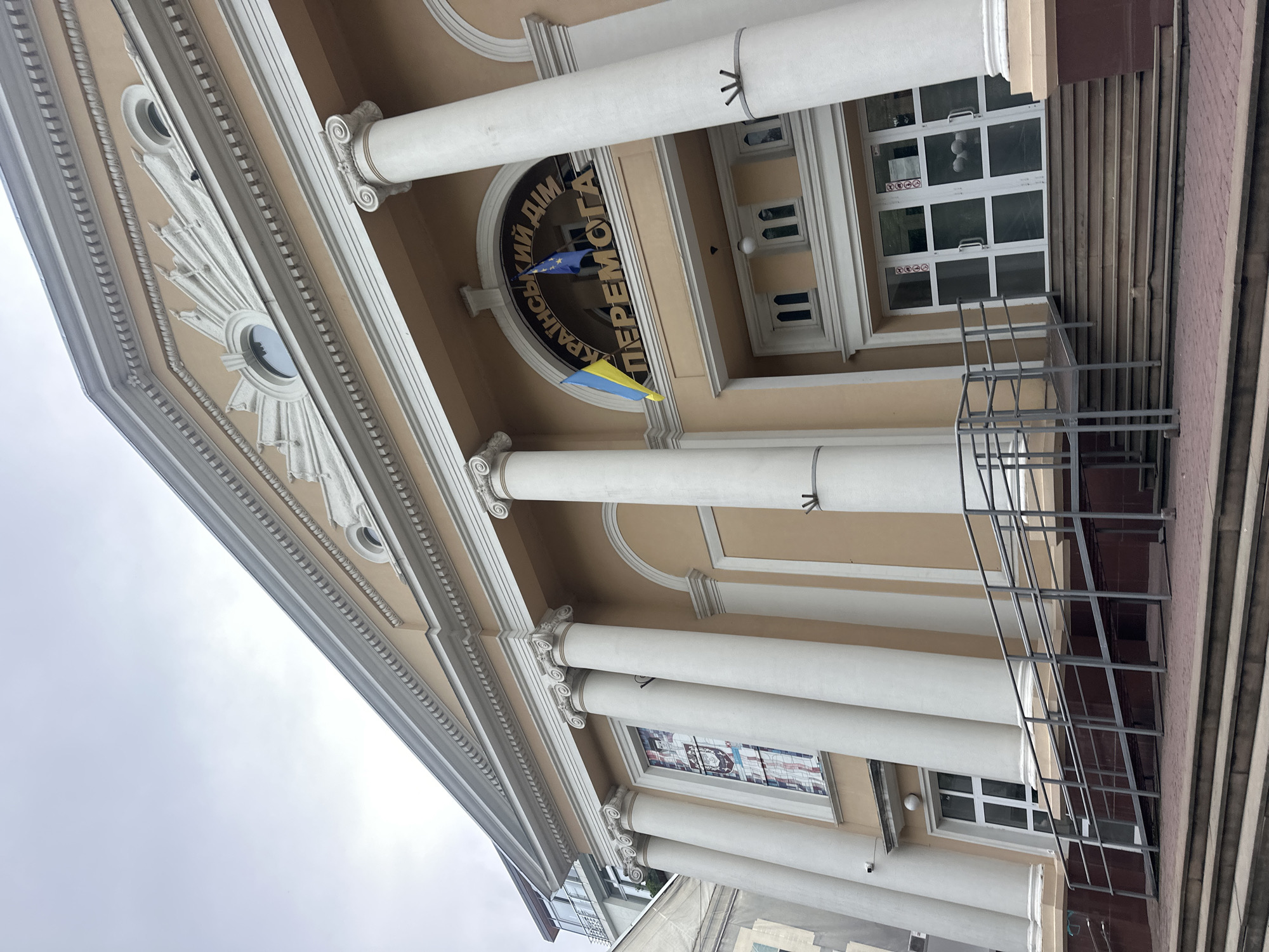 The image shows the front facade of the Ukrainian House "Perexmoga" in Ternopil, Ukraine. The building features classical architecture with large white Corinthian columns, a pediment with decorative elements, and a light peach-colored exterior. Two flags, the Ukrainian flag and the European Union flag, are displayed above the entrance. The entrance itself has large glass doors and is approached by a set of stairs with a metal ramp to the left. There is a stained-glass window visible to the left of the entrance. The sky is overcast.