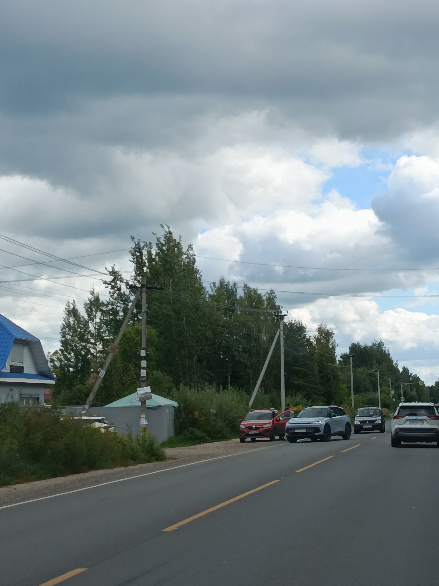 The image depicts a roadside scene on a cloudy day, likely in a rural or suburban area. A paved road with a yellow line down the center runs from the foreground to the background. Several cars are visible on the right side of the road, some moving and some parked. A white SUV is in the foreground on the right, followed by a dark-colored car, then a light blue crossover SUV, and a red hatchback with its driver's door open.
On the left side of the road, there is a house with a blue roof and white siding, surrounded by dense green foliage. Further down the road, a utility pole with wires is prominent, with several signs attached to it. A light gray, tent-like structure is situated behind the utility pole and to the left of the road. Trees form a dense line along the horizon behind the road and the house. The sky is filled with a mixture of dark and light clouds, with patches of blue sky visible.
