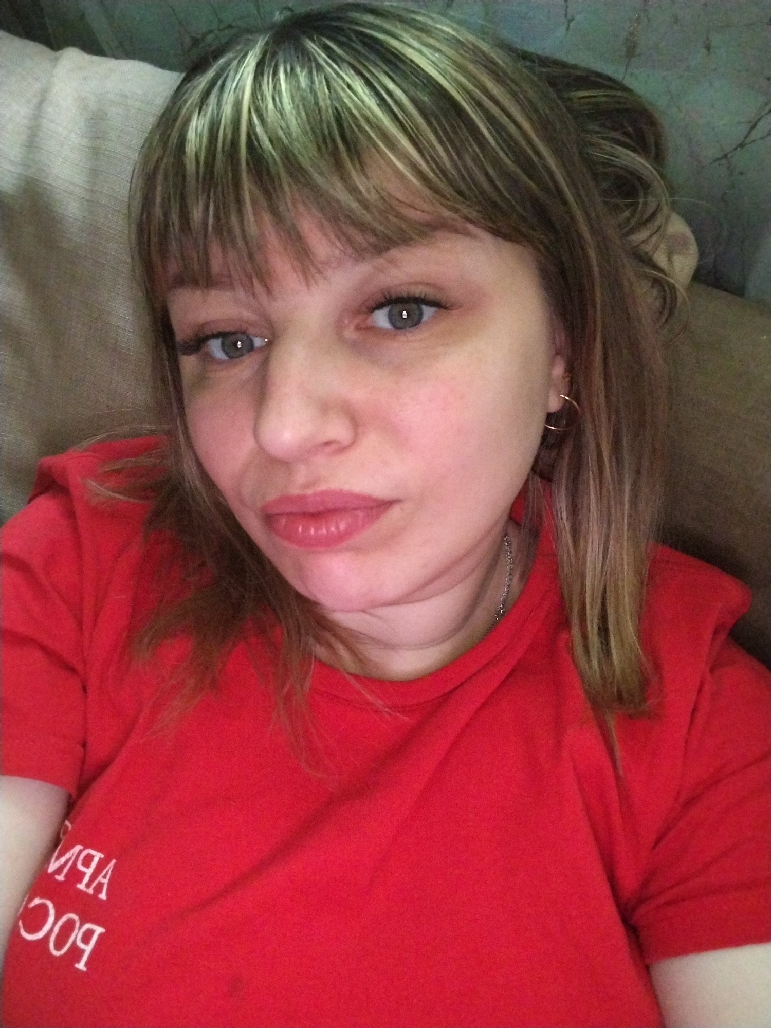 A woman with light skin, light-colored eyes, and shoulder-length blonde hair with bangs is lying down. She is wearing a red t-shirt. A silver necklace is visible around her neck, and she has gold hoop earrings. The background consists of a beige and green textured surface. The letters "РОССИЯ АПР" are visible in white on the t-shirt. The location is Заволжье, Russia.