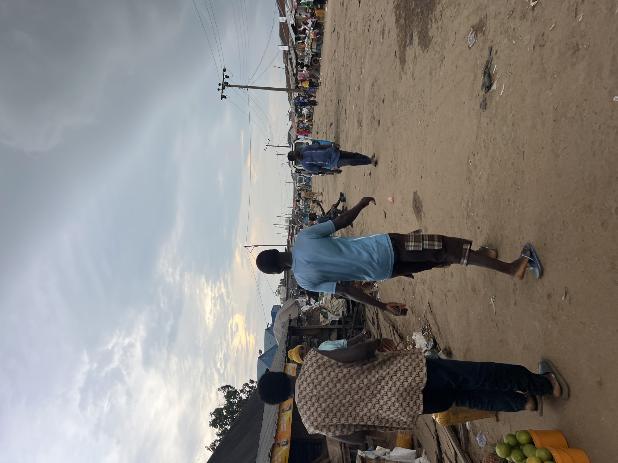 The image depicts a street scene in Oko Amakom, Nigeria. The ground is unpaved and dusty, with scattered debris. Several people are visible, walking and standing. In the foreground, two men are walking away from the camera. The man on the left is wearing a patterned sleeveless vest and dark pants. The man on the right is wearing a blue t-shirt and checkered shorts. Further down the street, other individuals and market stalls can be seen. Vehicles, including what appears to be a tricycle and a van, are present in the background. Power lines and poles are visible against a cloudy sky. The overall impression is that of a bustling outdoor market in a somewhat informal urban setting.