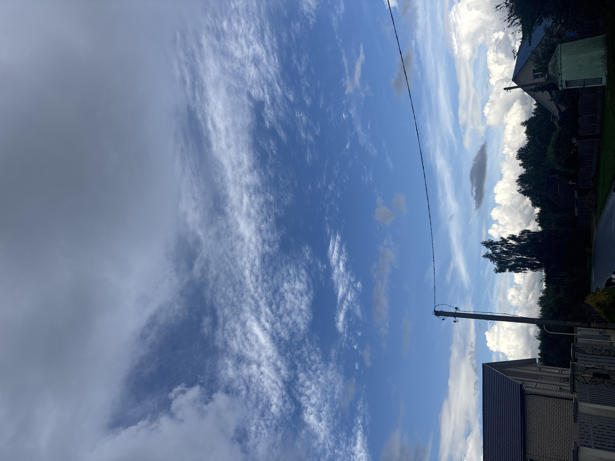 The image depicts a rural landscape under a partly cloudy sky. In the foreground, on the left, a brick house with a dark roof is visible, partially obscured by a fence. To the right of the house, a wooden utility pole stands tall with wires stretching across the frame. Beyond the pole, a dense line of dark green trees forms the horizon. In the middle ground, a paved road or path is visible, with what appears to be a puddle of water on its surface. Two residential buildings, one with a blue roof and another smaller structure, are nestled amongst the trees on the right side of the image. The sky is a mix of bright blue and scattered white clouds, with some wispy cirrus clouds higher up. The overall impression is one of a quiet, everyday scene in a rural settlement within the Капыльскі сельскі Савет region of Belarus.