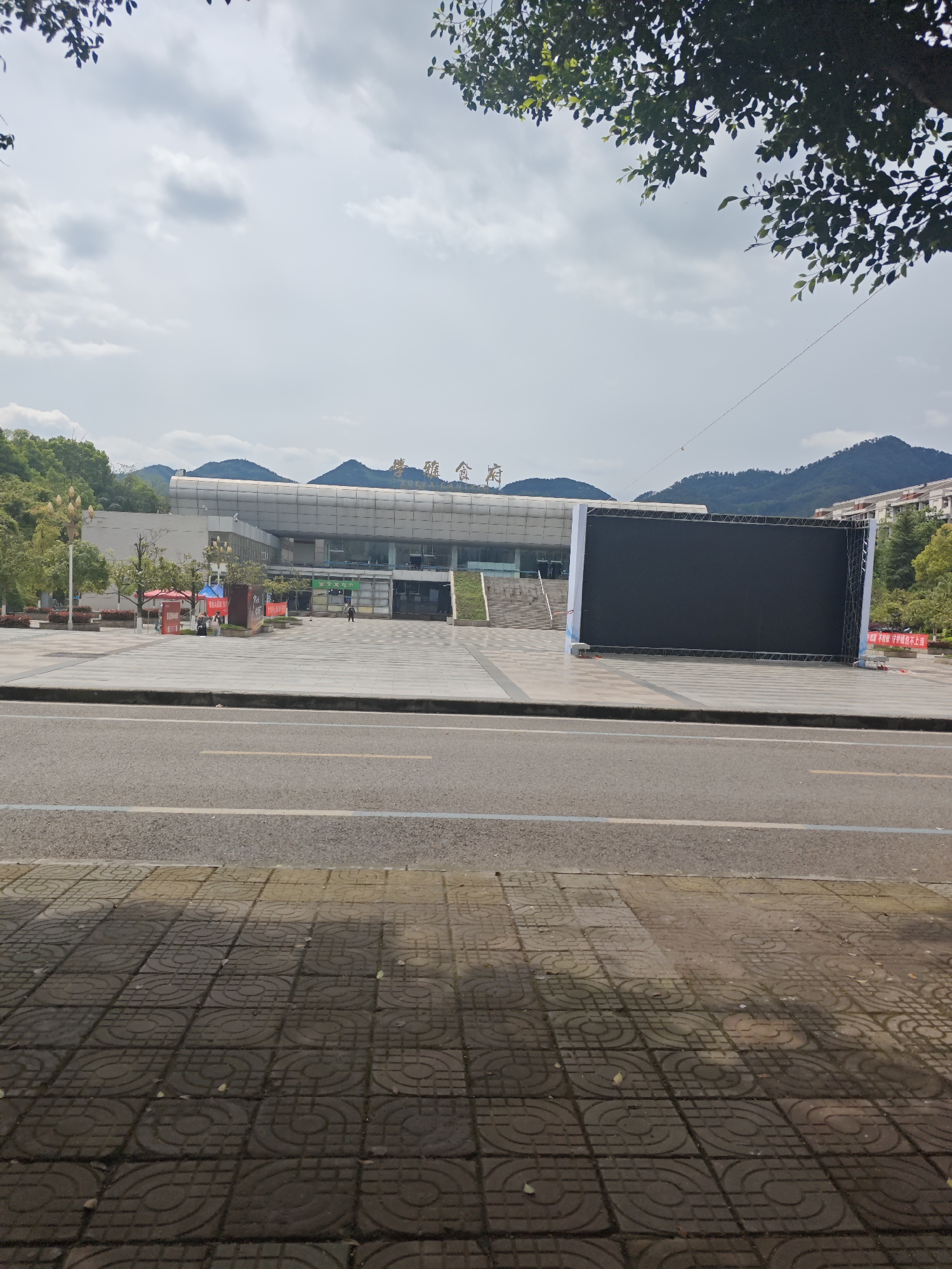 The image shows a large building identified as the "Xue Ya Cang" Innovation Center in the Chongqing High-tech Zone, China. The building has a modern architectural style with a combination of glass and concrete. In front of the building, there is a paved area, a road, and a large black screen set up on the right side of the building. Distant mountains are visible in the background. A few people are seen in the vicinity of the building. The sky is overcast with clouds. A tree with foliage is visible in the upper right corner. The foreground shows the pavement on which the photographer is standing.