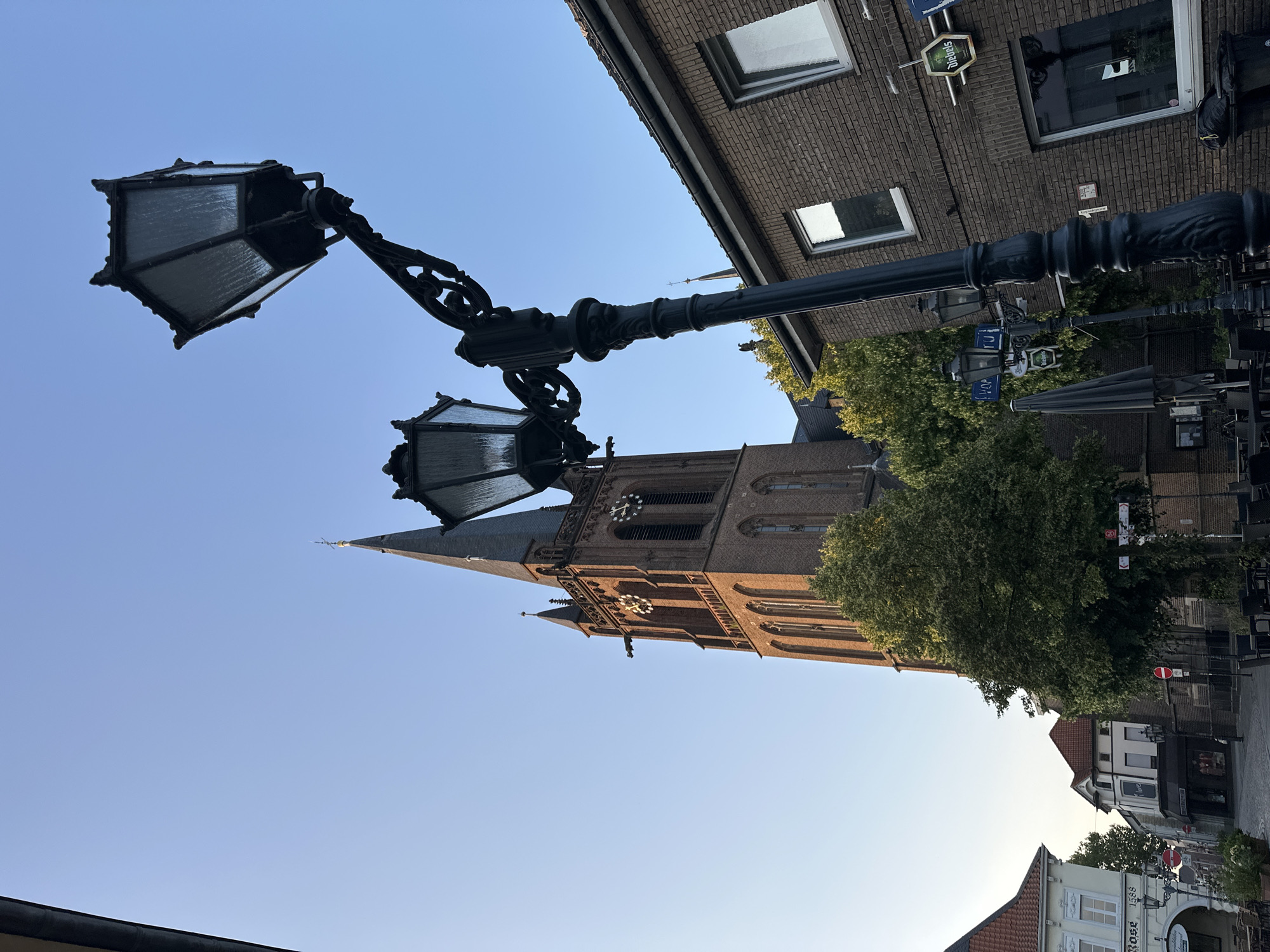 This is a vertical, eye-level, outdoor, full shot of a street scene in Krefeld, Germany, on a clear, sunny day with a blue sky. A tall, gothic-style brick church with a spire dominates the background on the left. In the foreground, a black, ornate lamppost with two lanterns extends diagonally from the bottom right to the top center. To the right of the lamppost is a brick building with windows. Trees are visible between the church and the lamppost. Signs and more buildings are visible in the distance on the left side of the street. The scene appears to be in a pedestrian area with outdoor seating for what appears to be a cafe or restaurant. There are no people visible in the frame.