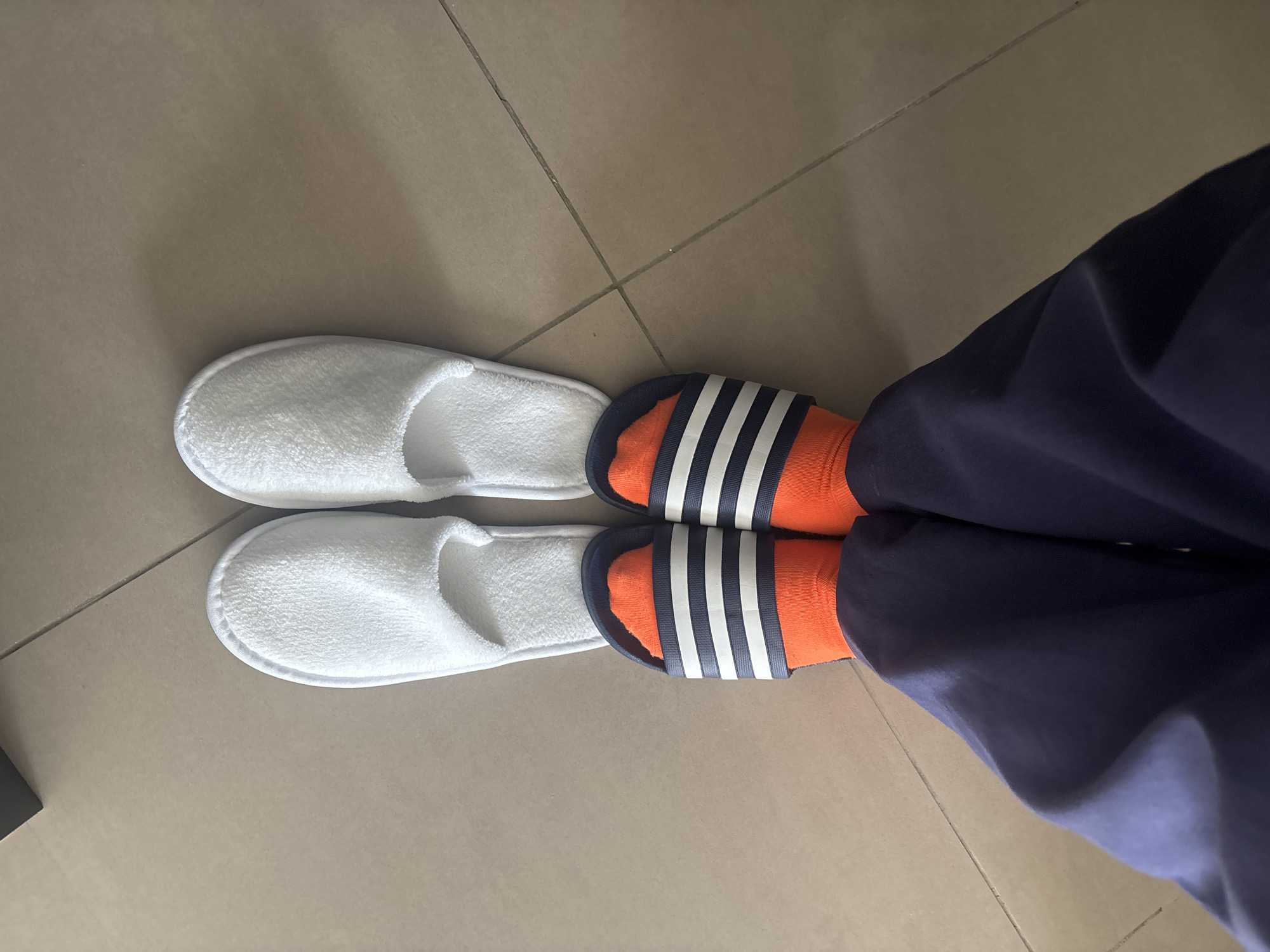 A top-down shot shows a person standing on a tiled floor. They are wearing dark blue pants, bright orange socks, and a pair of dark blue slides with white stripes. Behind them, there are two white terry cloth slippers. The floor is made of square beige tiles. The scene appears to be indoors, possibly in a hotel room in Dubai, United Arab Emirates, given the style of slippers.
