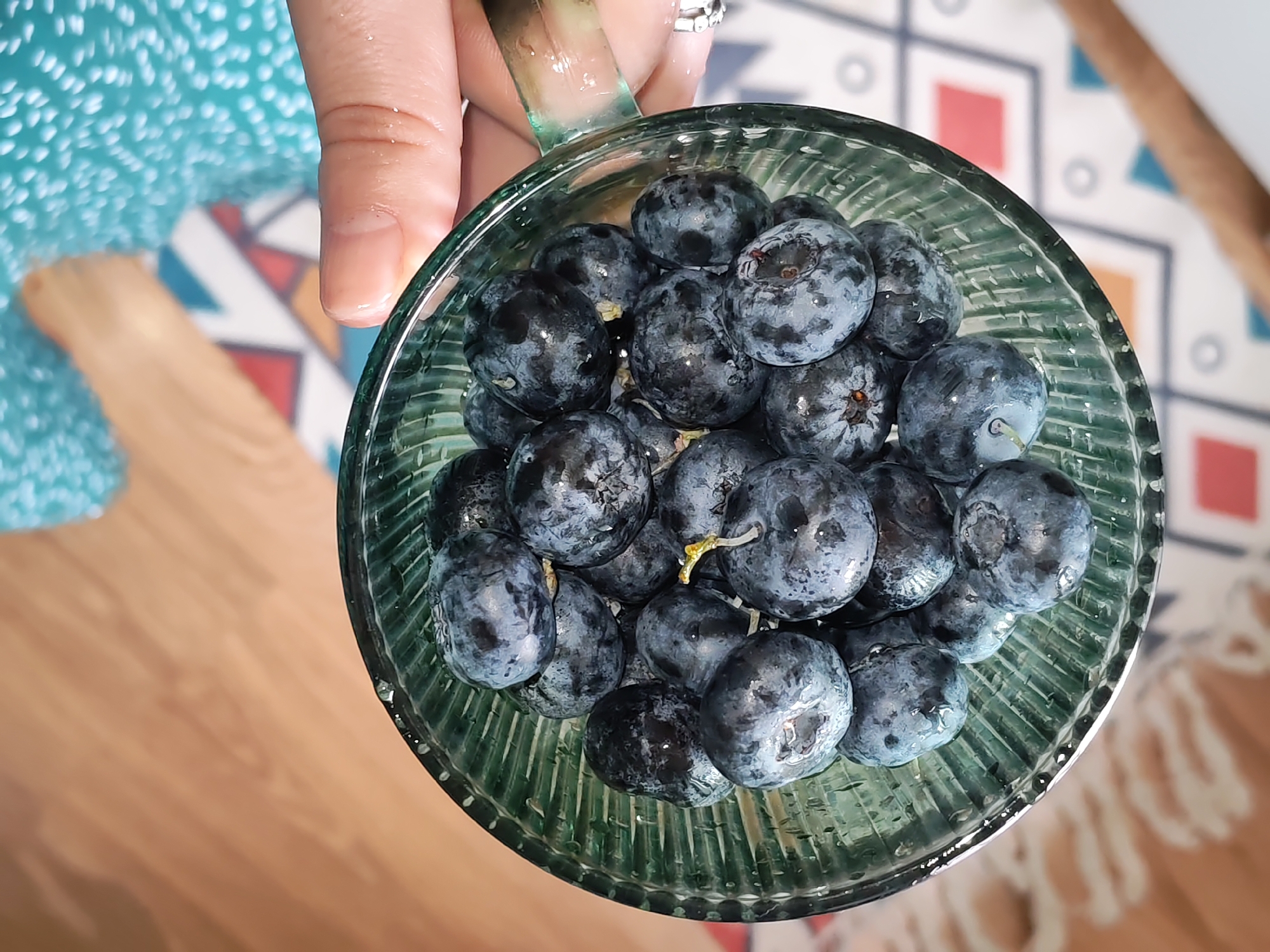 The image captures an overhead view of a person holding a glass filled with fresh blueberries. A light-skinned hand, adorned with a silver-toned ring on the ring finger, holds a clear, green-tinted glass mug or bowl. The glass has a ribbed or fluted design. Inside, numerous dark blue blueberries fill the container, many displaying a natural powdery 'bloom' and small water droplets, suggesting they are freshly washed. Some blueberries still have tiny green or yellow stems attached.
In the blurred background, part of a person's leg or foot is visible, covered by a light blue fabric with a textured or patterned surface. A wooden floor extends beneath. Further back, a colorful geometric patterned rug or mat, featuring shapes in red, blue, orange, and white, is visible on the floor. The scene depicts an indoor setting, likely a home. There are no discernible time-of-day indicators, specific weather conditions, or visible text. The focus is on the ripe fruit being held.