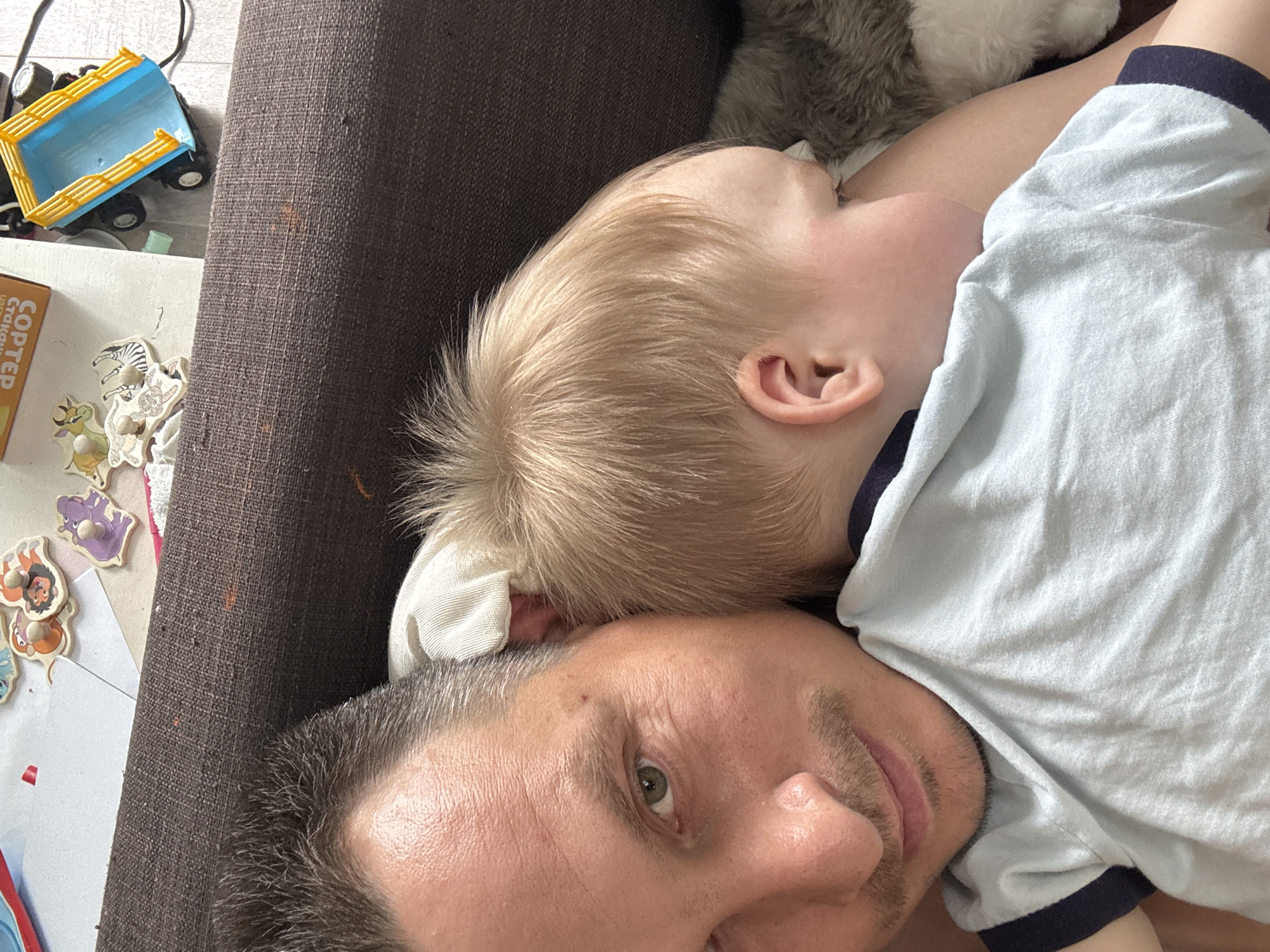 A man and a child are lying down, possibly on a couch. The man's face is visible, and he is looking toward the camera. The child, with blond hair, is lying on the man. The setting is indoors, next to a side table with toys and a box. The location in Izhevsk, Russia cannot be determined from the image.
