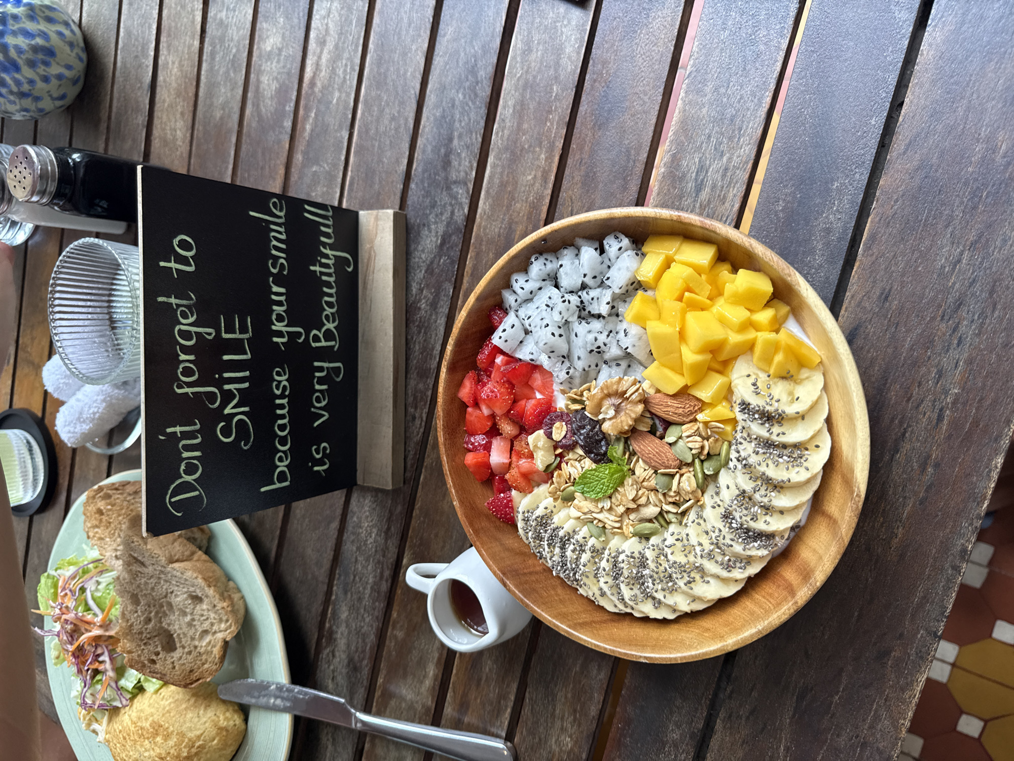 The image shows a wooden table setting in a cafe in Đà Nẵng, Vietnam. A wooden bowl filled with fruit, granola, and chia seeds dominates the scene. The bowl contains sections of sliced bananas topped with chia seeds, cubed mango, white dragon fruit, and diced strawberries. A small white cup containing a dark liquid, possibly syrup, sits adjacent to the bowl. A black sign with white lettering that reads "Don't forget to SMILE because your smile is very Beautiful!" stands upright, next to a plate of a sandwich or open-faced sandwich with salad and a pastry. Silverware (a knife) is placed next to the plate. Other items visible include a glass, condiments, and an additional dish, all arranged on the wooden table. The table shows wear and tear, with various scratches and imperfections, suggesting it is well-used. The image is taken from a slightly elevated perspective, and the light appears to be natural.