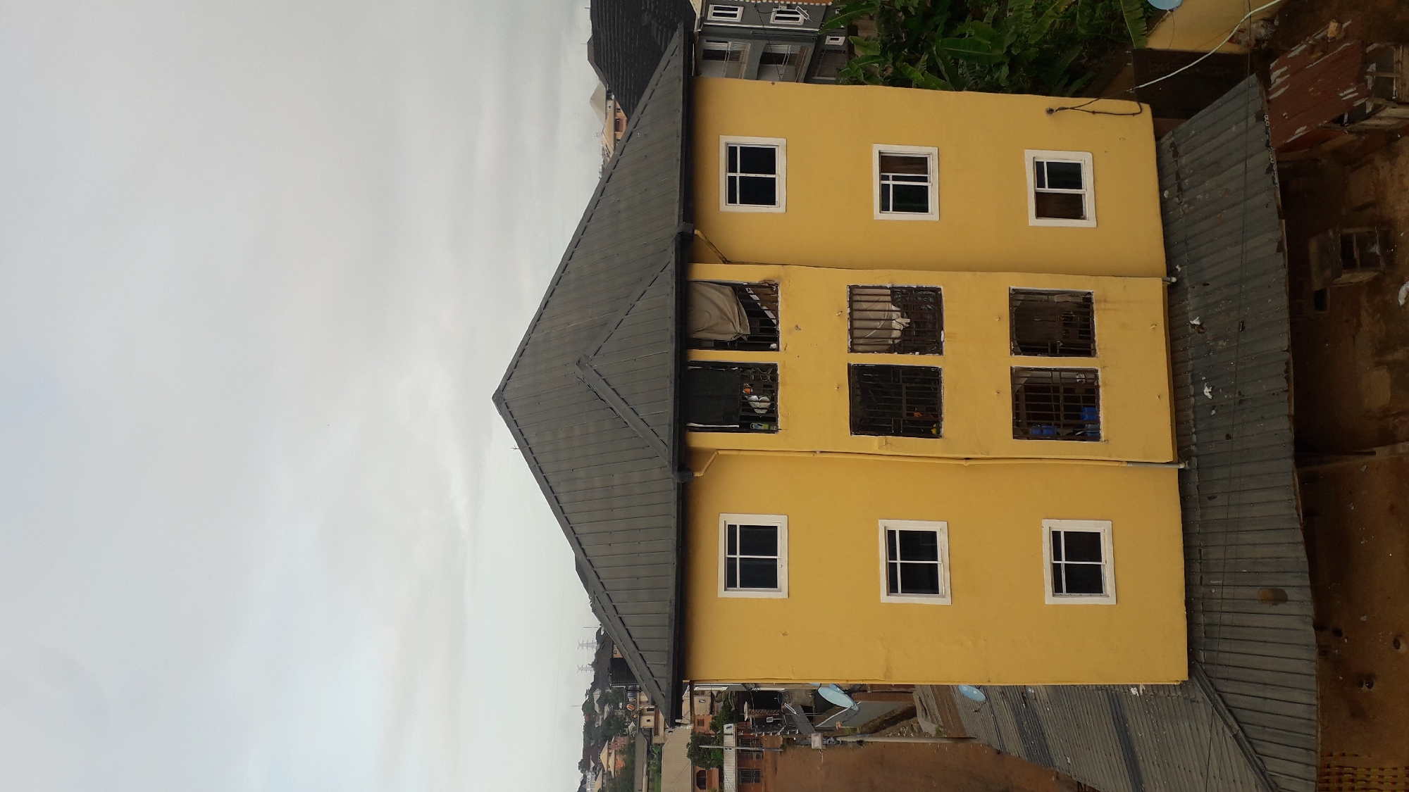 The image depicts a yellow, two-story building with a dark gray, gabled roof in Nkpor, Nigeria. The building has a rectangular shape, composed of two adjacent modules sharing the roof. It is viewed from an elevated perspective. Each module has three windows per floor. The ground floor and top floor windows are simple squares. The windows on the middle floor have metal security bars. The surrounding environment includes other buildings, rooftops, and vegetation. The sky is overcast.