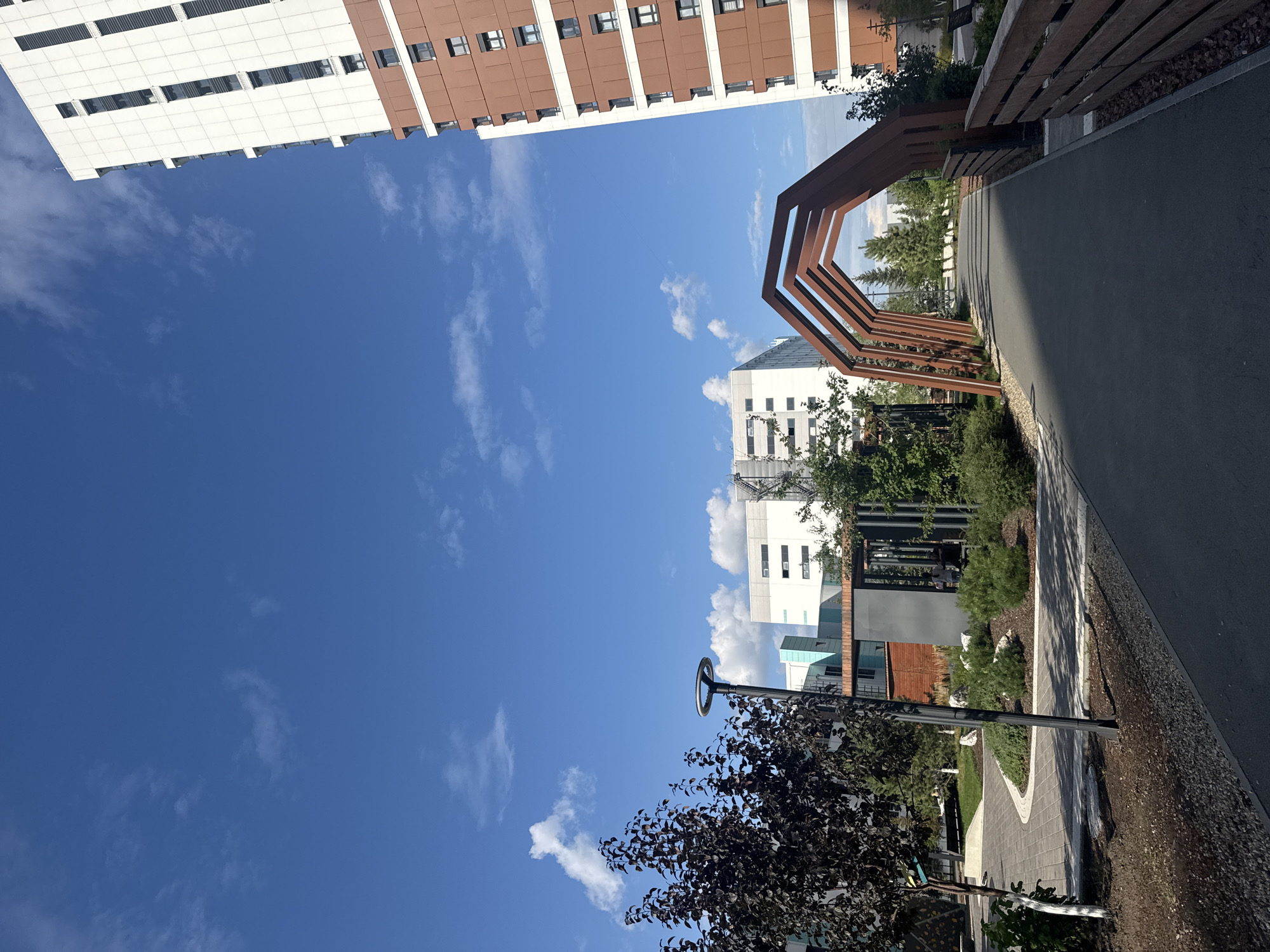 An outdoor scene on a clear day shows a modern urban courtyard. In the foreground, a paved pathway leads through a landscaped area. To the right, a tall, multi-story building with a brown and white facade dominates the right side of the frame. To the left, a dark-leafed tree stands next to a lamppost, with more greenery and modern buildings in the background. A distinctive wooden archway structure spans the pathway. The sky is a bright blue with scattered white clouds.
The scene is located in the "Zolotaya Dolina" residential complex in Novosibirsk, Russia.