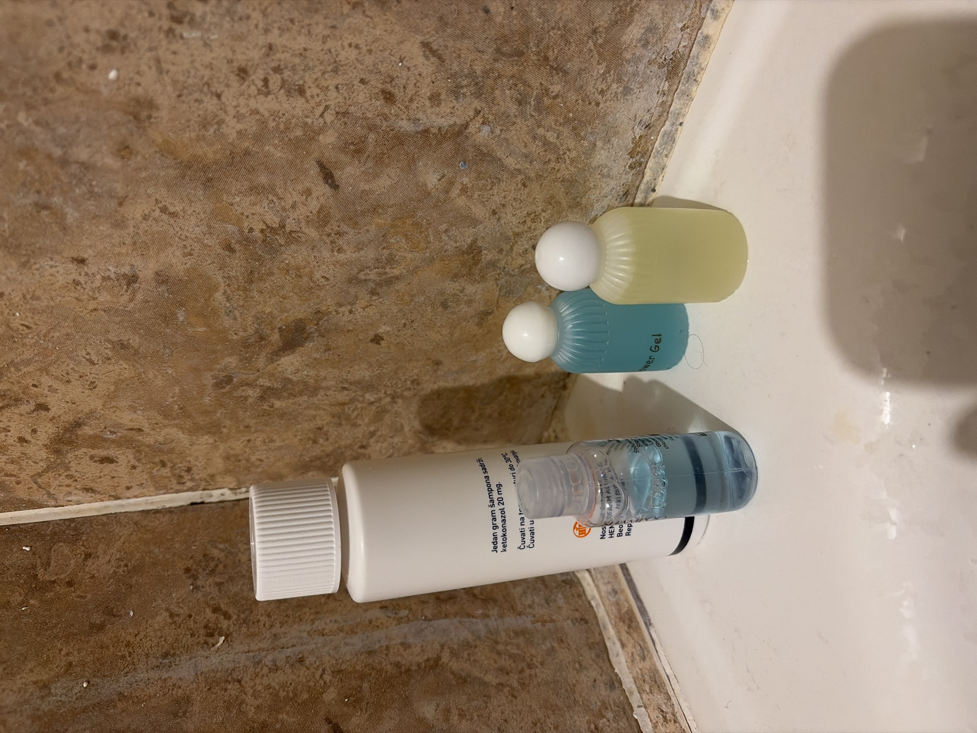 A top-down, close-up shot depicts a bathroom scene, possibly in a hotel or apartment in Budva, Montenegro. The image features three small bottles or containers placed on what appears to be a tiled wall or the edge of a bathtub. The surface is stained with water and cleaning product residues.
The largest container is white with a ribbed cap and has a clear, multi-chambered section filled with a blue liquid. Text on the container indicates the presence of "ketokonazol 20 mg" per gram of shampoo, suggesting it's a medicated shampoo. The label also includes storage instructions in Serbian, indicating that it should be stored at temperatures up to 30°C. The brand name appears to be "HEN Beofarm Altr".
The other two containers are smaller, capsule-shaped bottles with white caps. One contains a yellow liquid, while the other contains a blue liquid. One is labelled "ower Gel".
The location is inferred by the text present on the bottle.