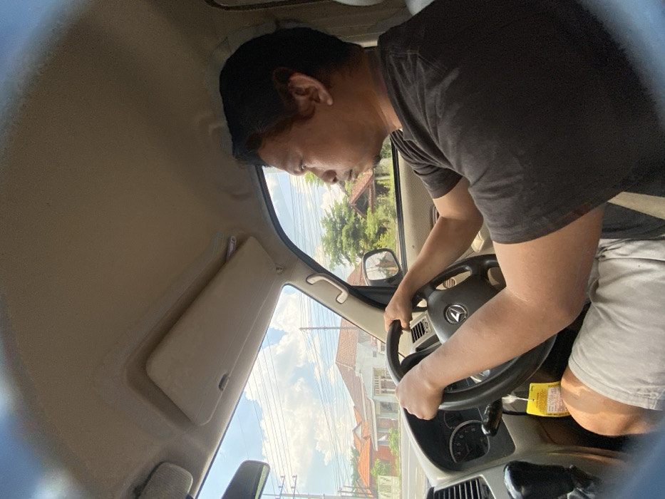 A man in a black t-shirt drives a car with his hands firmly on the steering wheel. He appears focused, looking down slightly as if concentrating on the road. The interior of the vehicle is visible, including the dashboard, sun visor, and window. Through the window, a view of houses and trees under a partly cloudy sky can be seen. The image is taken from the passenger seat.