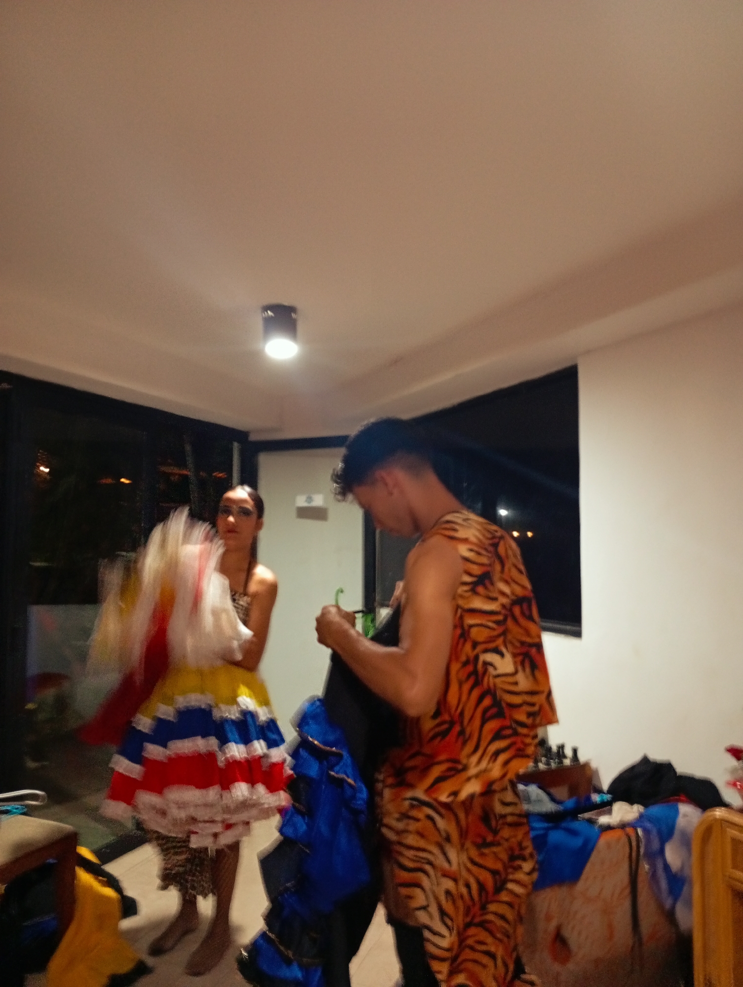 The image shows a room in Playa El Agua, Venezuela, where two people are dressing in costumes. The room has light-colored walls and a single light fixture in the center of the ceiling. A woman is on the left side, wearing a dress with white, yellow, red, and blue layers. She seems to be adjusting the dress. On the right, a man is dressed in a tiger-print outfit. He appears to be holding a garment bag. Various blue and black items are scattered on a surface behind him, along with some personal items. A window is visible in the background, with lights visible outside, suggesting it is nighttime.