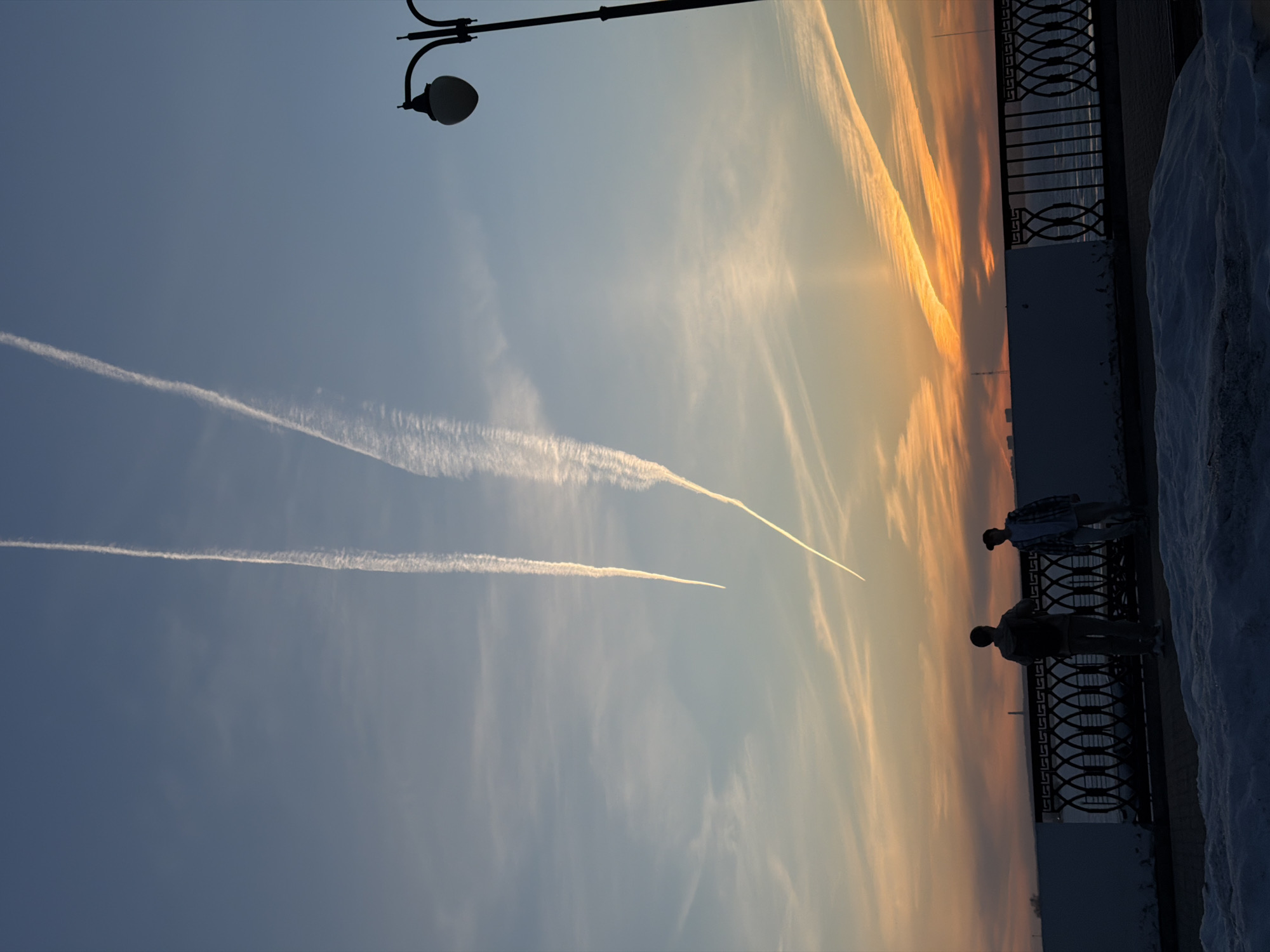 The image depicts a sunset over the city of Izhevsk, Russia. In the foreground, two individuals stand near a wrought-iron railing overlooking a body of water, potentially the Izhevsk Pond. Patches of snow are visible along the side of the railing. A street lamp is situated nearby. The sky displays contrails from aircraft against the backdrop of the sunset. The horizon shows orange and yellow hues.