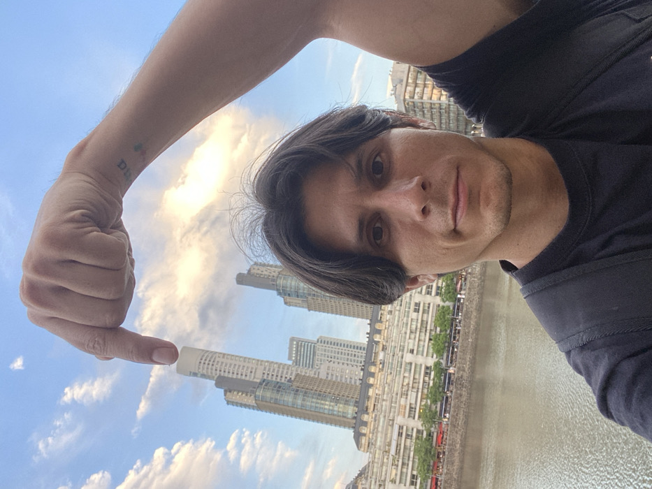 A man in a black shirt points to a tall building with his finger. The building has a white roof. The man has short brown hair and brown eyes. There is a river and other buildings in the background. The sky is blue with white clouds. A tattoo is visible on the man's arm. The picture is rotated counterclockwise.