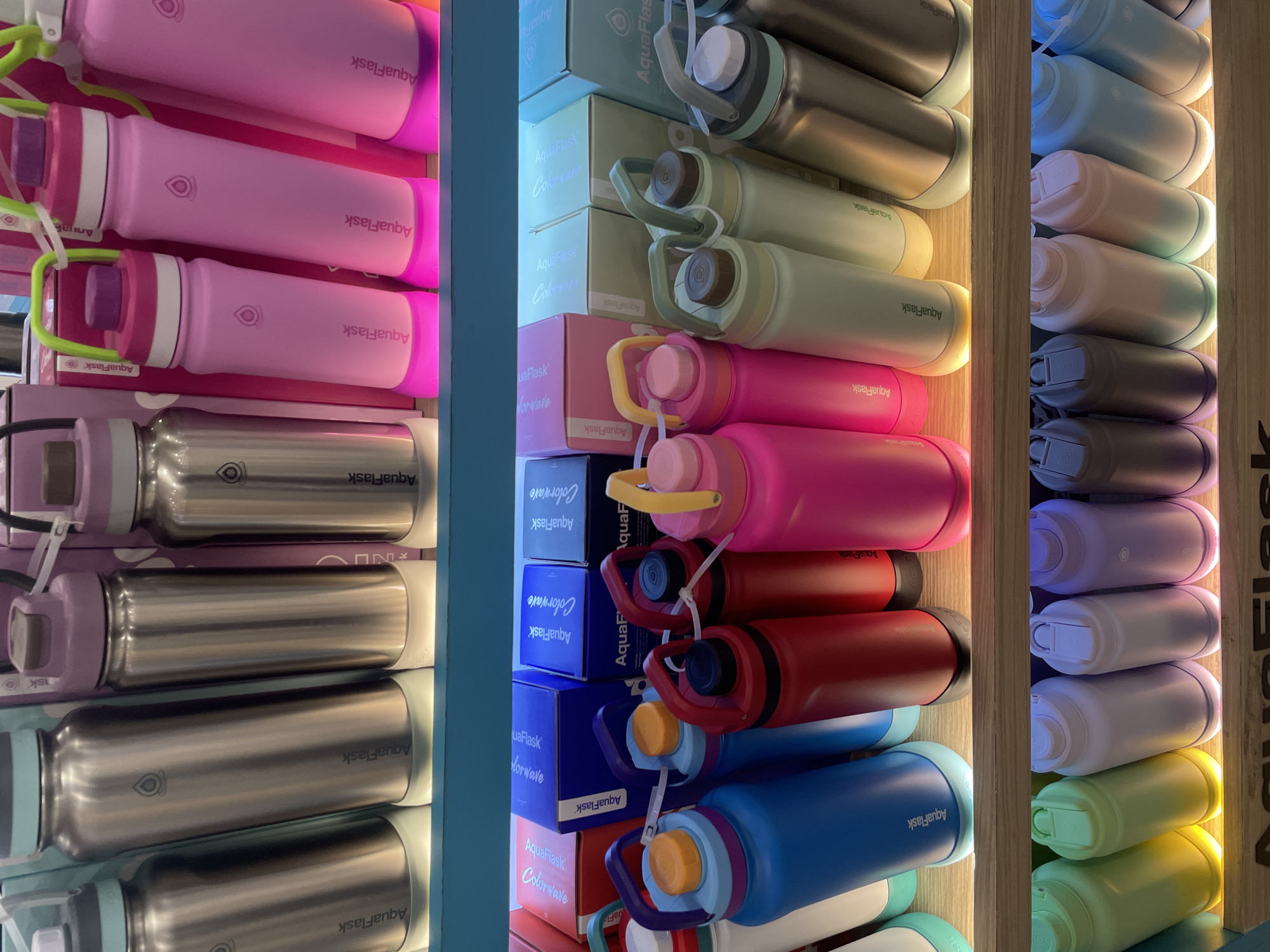 The image displays a retail environment, likely a store, featuring an extensive collection of AquaFlask brand water bottles. These bottles are arranged on wooden shelves, illuminated from below by soft, colored lights, creating a visually appealing display. The bottles vary in color and size, with some of the larger ones visible on the top shelf in metallic silver and pink hues. The middle and bottom shelves showcase a wider spectrum of colors including blue, red, pink, light green, and white. Some bottles have prominent handles, suggesting they are designed for portability. Boxes with the AquaFlask branding are also visible behind some of the bottles on the middle shelf. The overall impression is one of an organized and well-stocked merchandise display. This scene is located at the AquaFlask store in SM Lanang Premier, Davao City, Philippines. There are no people visible in the image.
