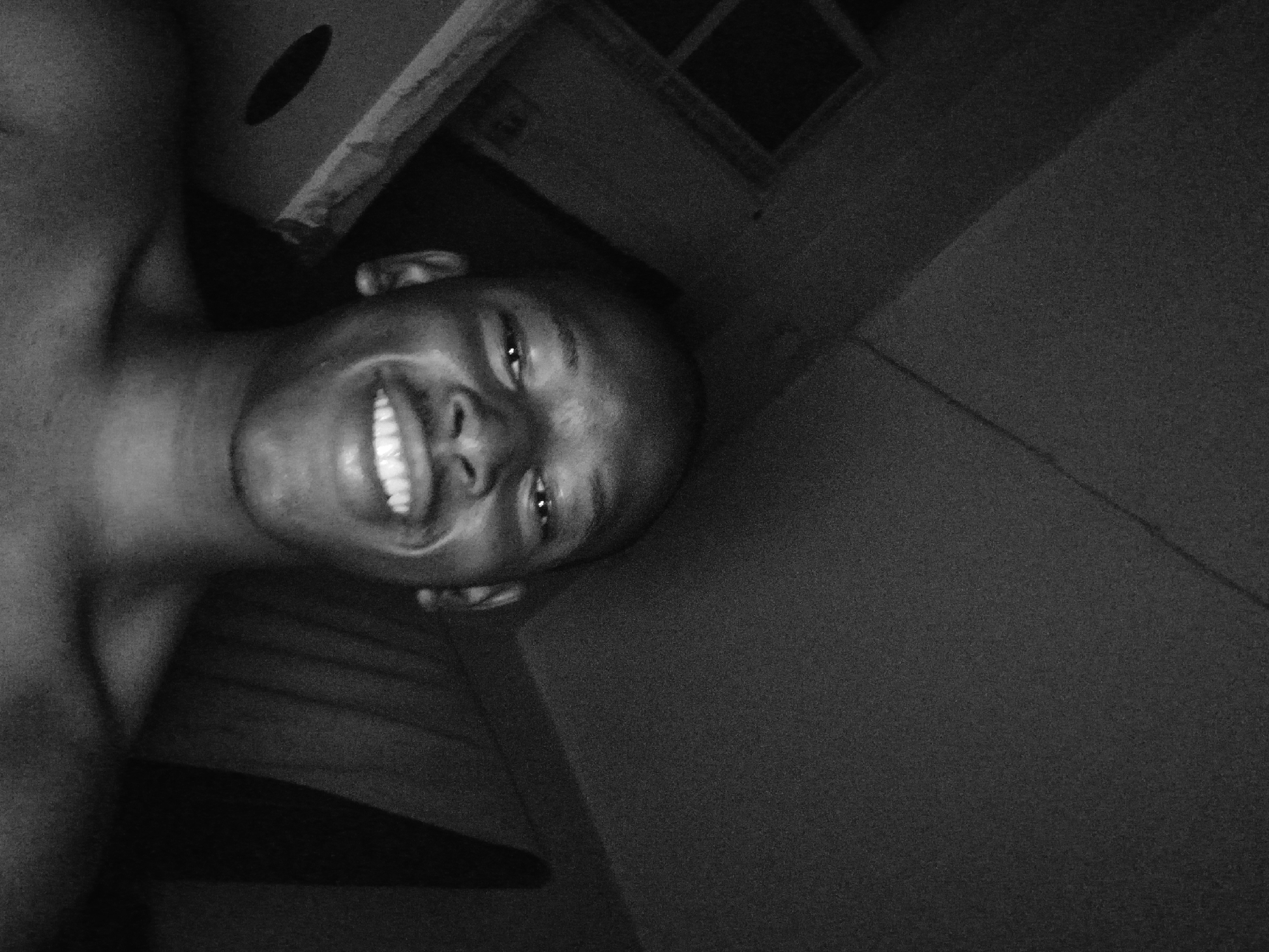 A black and white, low-light, close-up selfie of a young Black male. He is shirtless and looking directly at the camera with a wide smile, showing his teeth. His head is bald, and he has a small mustache. The background is dark and out of focus, appearing to be an indoor setting with some shadows suggesting a ceiling and wall. There is a hint of some object or structure to the left of the subject. The image quality is grainy, typical of low-light photography. The location is not discernible from the visual information provided in the image.